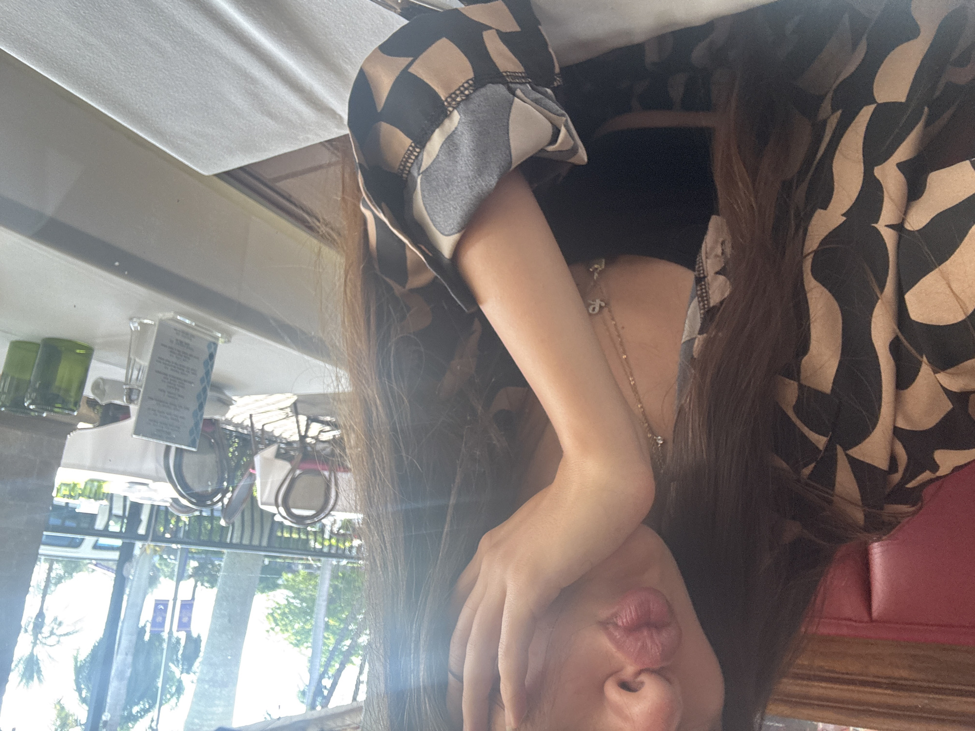 A woman sits at a restaurant table in Boca Raton, Florida. She wears a brown, black, and beige patterned shirt. She holds her left hand up, partially covering her face, and pouts her lips. In the background, trees, buildings, and a bus are visible through a glass wall. Table settings and outdoor seating are also visible in the background. The table she sits at is covered with a white cloth. A red padded seat is visible on the right.