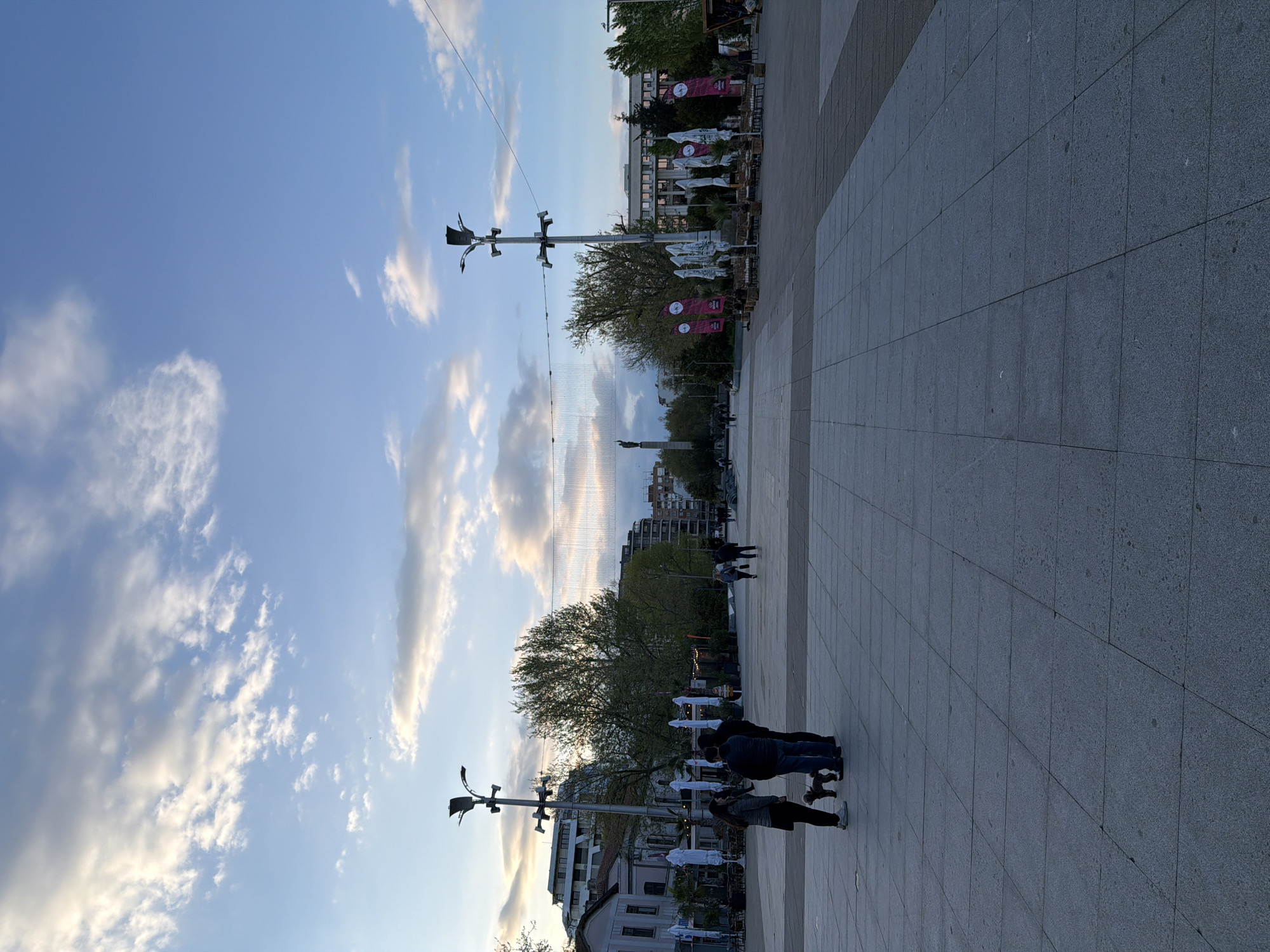 The image captures a plaza in an unknown city, with a paved ground leading towards the distant buildings and a statue. The sky is visible with scattered clouds. Two distinct light poles stand on either side of the image. People are walking around the plaza. A row of white umbrellas and red banners suggests a cafe or an outdoor establishment. The image is taken from a low angle.