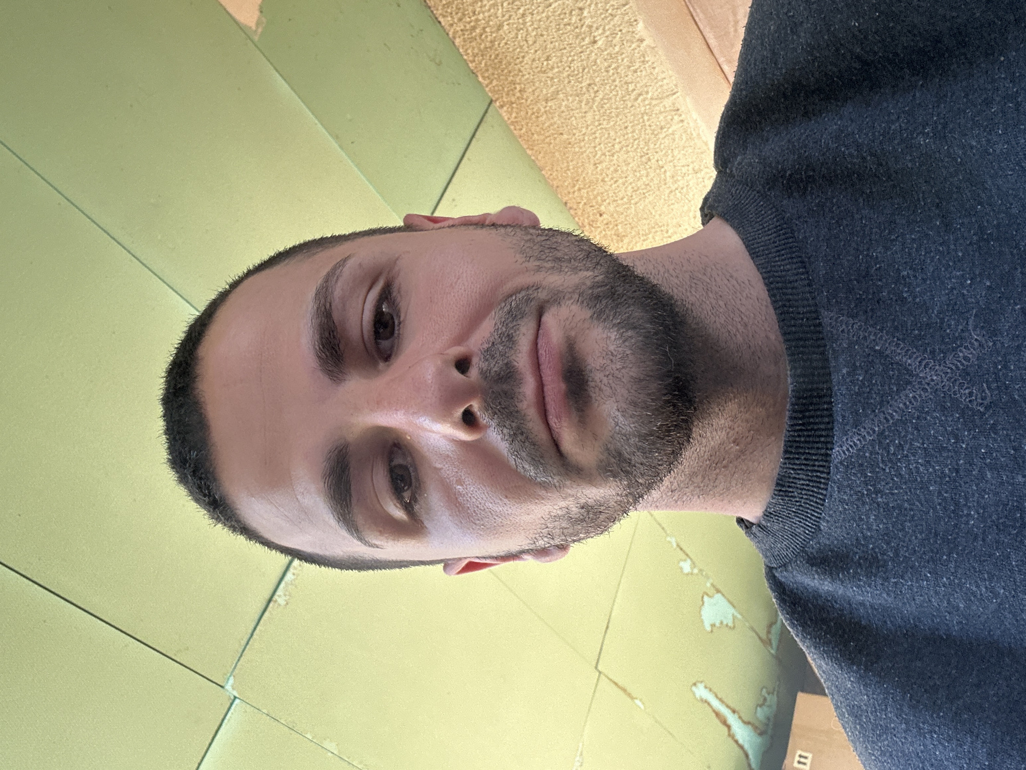 A close-up self-portrait of a man with short dark hair, a stubble beard, and brown eyes. He is wearing a dark gray sweatshirt. The background appears to be a pale green tiled wall. The image is rotated clockwise. Location details are not present in the image.
