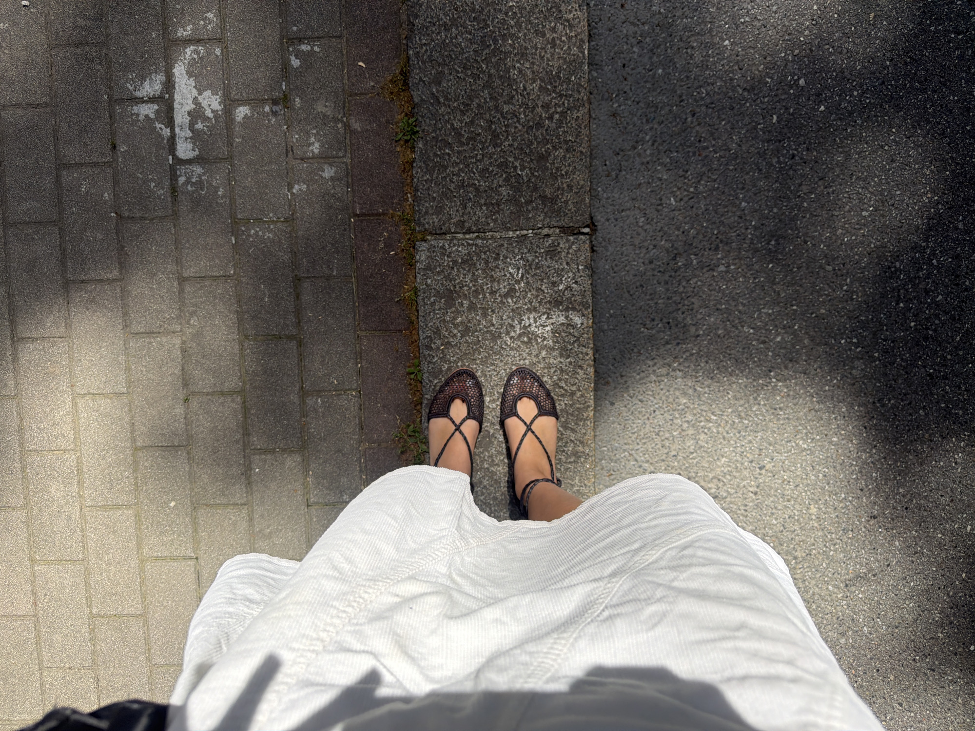 The image is a high-angle shot of a person standing on a street in Milan, Italy. The person is wearing a long white corduroy skirt and brown lace-up sandals. Their feet are positioned on the edge of a curb separating a sidewalk made of concrete pavers from a paved road. The sidewalk has some discolored patches and small vegetation growing in the cracks. The road is made of asphalt. The lighting suggests a sunny day.
