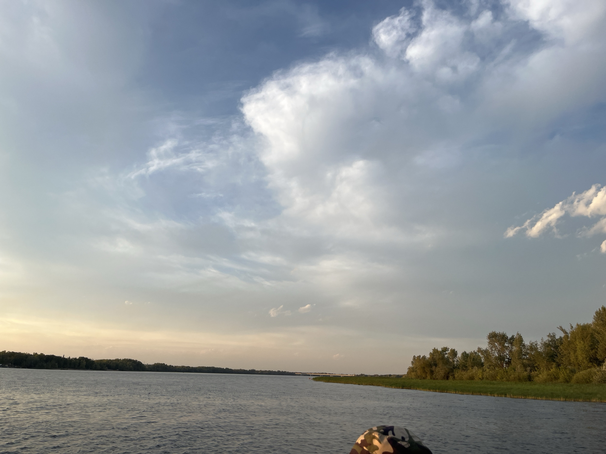 The image depicts a wide river, possibly the river Oka, near the village of Shumeika, Russia. The sky is filled with a mix of wispy and cumulus clouds, with a hint of sunset colors on the horizon. The river's surface is rippled, indicating a gentle breeze. On the right bank, a dense line of green trees and reeds borders the water. In the foreground, partially visible at the bottom right, is a person wearing a camouflage-patterned hat, suggesting they are on a boat or at the river's edge. The overall scene conveys a sense of tranquility and natural beauty.