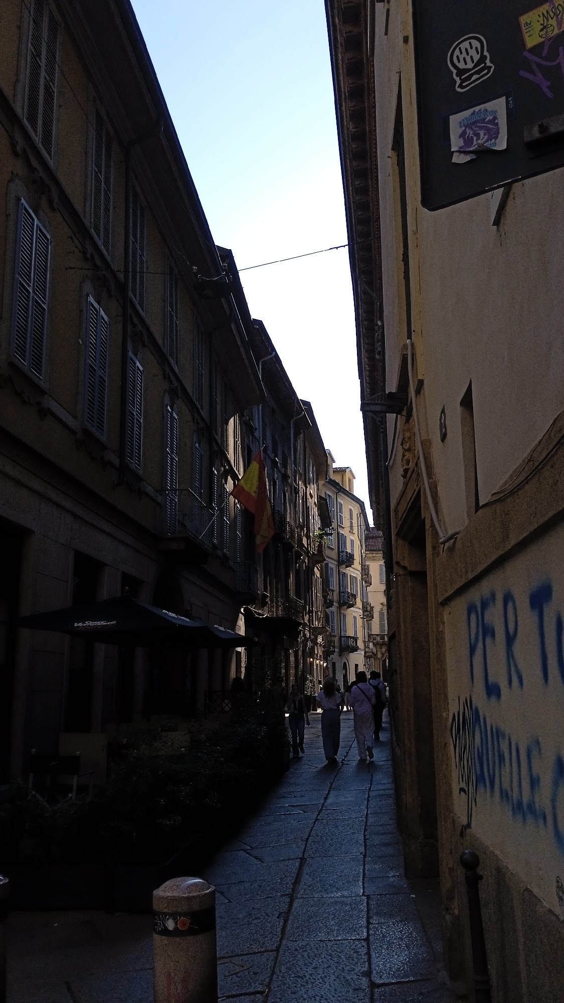 The image depicts a narrow street in Milan, Italy, with tall buildings lining both sides. The street is paved with stone and is partially shaded. On the left, a building with multiple stories features closed shutters and balconies. A yellow and red flag is visible hanging from one of the balconies. To the right of the street, a building wall is covered in graffiti. Several people are walking away from the camera down the street. In the foreground, there is a bollard with some decorative elements. The sky above is clear and blue.