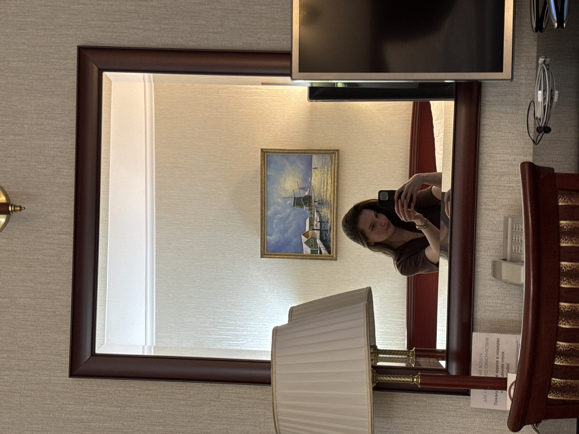 A woman is taking a mirror selfie in a room. She is holding a black smartphone in her right hand, angled towards herself. She is wearing a brown v-neck shirt. In the reflection of the mirror, a bed with white bedding is visible behind her. To the left of the mirror, a cream-colored lampshade with a gold trim is visible. Above the bed, a framed painting of a windmill by a body of water is hanging on the wall. To the right of the painting, a portion of a black television screen is visible. The room appears to be a hotel room, with textured beige wallpaper on the walls. A wooden desk is in the foreground, with a dark wooden chair partially visible. There is a small white sign with text in Russian on the desk. The overall lighting is soft and warm. The location is a hotel room in the city of Zhuralyovo (Kemerovo Oblast), Russia.