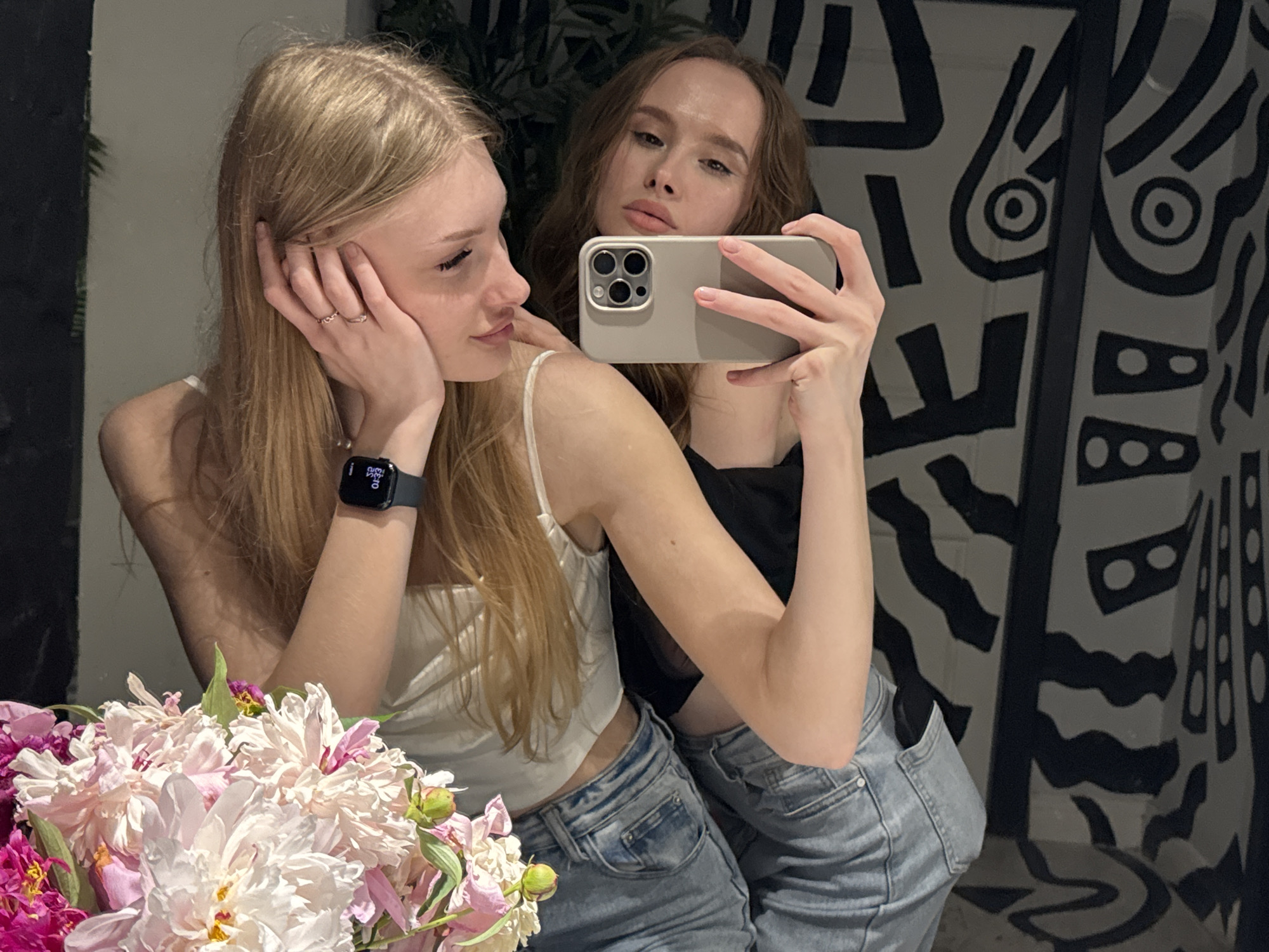This media file captures a selfie of two young women, likely in their late teens or early twenties, posing in what appears to be a residential setting, possibly an apartment in Perm, Russia. The woman in the foreground, with long blonde hair, is leaning her head on her hand, adorned with two rings on her fingers and a smartwatch on her left wrist. She wears a white camisole and light blue denim shorts. To her right, a woman with brown hair is also visible, holding a smartphone to take the picture. She is wearing a black top and similar denim shorts.
The background features a wall with a striking black and white abstract pattern, reminiscent of contemporary art or wallpaper. In the lower left corner of the frame, a generous bouquet of peonies in shades of pink and white adds a touch of vibrancy and floral elegance to the scene. The lighting suggests it might be daytime, possibly late afternoon, given the soft illumination. The overall mood is casual and friendly, with the subjects appearing relaxed and comfortable with each other. There is no visible text in the media file.