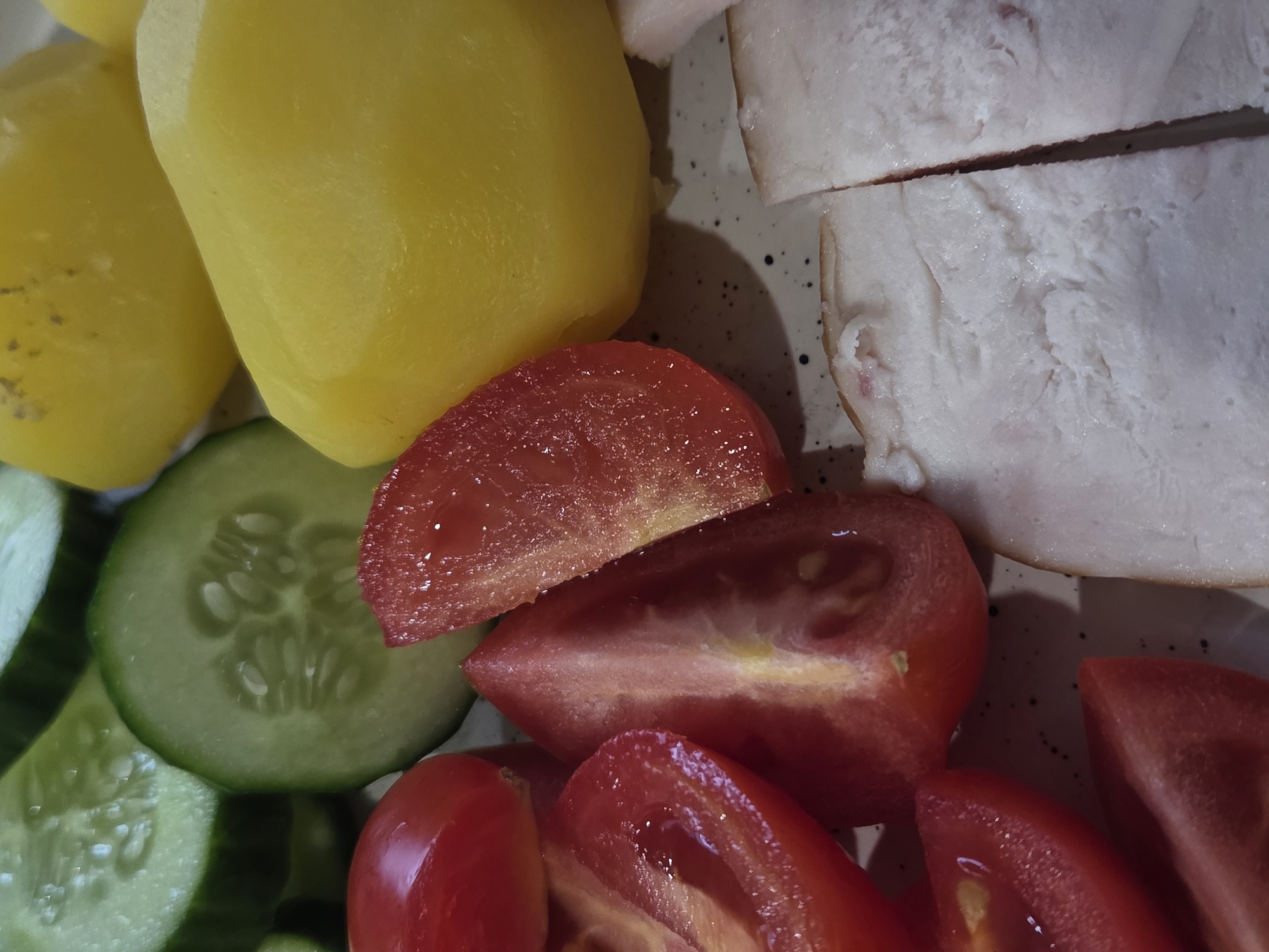 The image shows a close-up of a plate containing a variety of foods. These include boiled potatoes, sliced cucumbers, tomato wedges, and sliced bacon. The food appears to be arranged directly on the plate. The scene appears to be indoors.