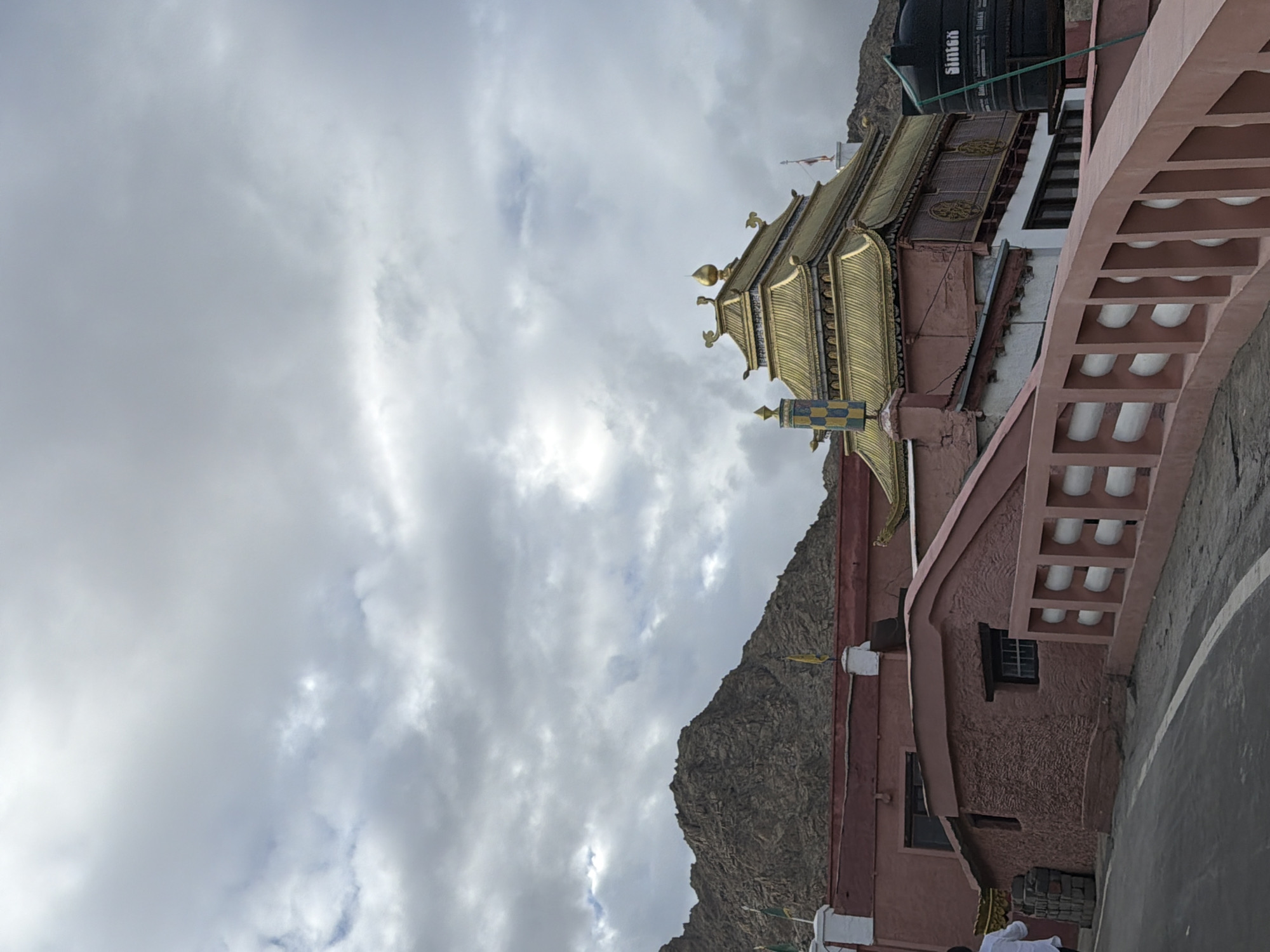 The image depicts a Buddhist monastery in Leh, India, under a cloudy sky. A prominent golden-roofed building, likely a temple or stupa, is visible. The monastery complex features pink-colored structures with traditional architectural elements. In the foreground, a road curves upwards, bordered by a pink fence with white circular designs. To the right, a large black water tank is installed on a rooftop. Mountains form the backdrop, adding to the scenic mountain landscape of Leh. A person in a white shirt is partially visible on the left, walking away from the camera.