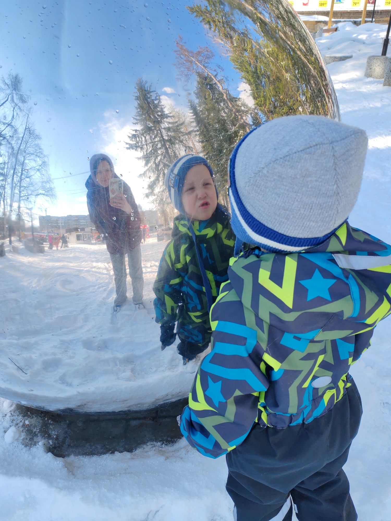 This outdoor image from Ust-Ilimsk, Russia, captures a vibrant winter scene centered around a child interacting with a large, curved, reflective mirror. The photograph is taken from behind a young child, who is wearing a gray and blue striped beanie, a brightly patterned blue, green, and black winter jacket adorned with star motifs, and dark snow pants. The child is engrossed, looking directly into their distorted reflection on the polished surface.
The reflection prominently shows the child's face with a concentrated or slightly questioning expression. Standing further back and to the left within the reflection is an adult, presumably a parent, dressed in a dark jacket, light pants, and a dark hood or beanie. The adult is holding a smartphone, appearing to take a photo or video of the child and the unique reflection.
The setting is a snowy public space, possibly a park or town square, bathed in bright daylight under a clear blue sky with some scattered clouds. Both bare deciduous trees and tall coniferous trees are visible in the background and are extensively reflected on the curved mirror. The ground is covered in a pristine layer of snow. Water droplets or ice remnants are visible on the mirror's surface, indicating cold temperatures. In the upper right of the reflection, distorted elements of the surroundings, including distant buildings and price signs (e.g., "189-60", "79-00", "119-00"), are visible, suggesting nearby commercial establishments. The image conveys a playful and curious moment during a winter outing.