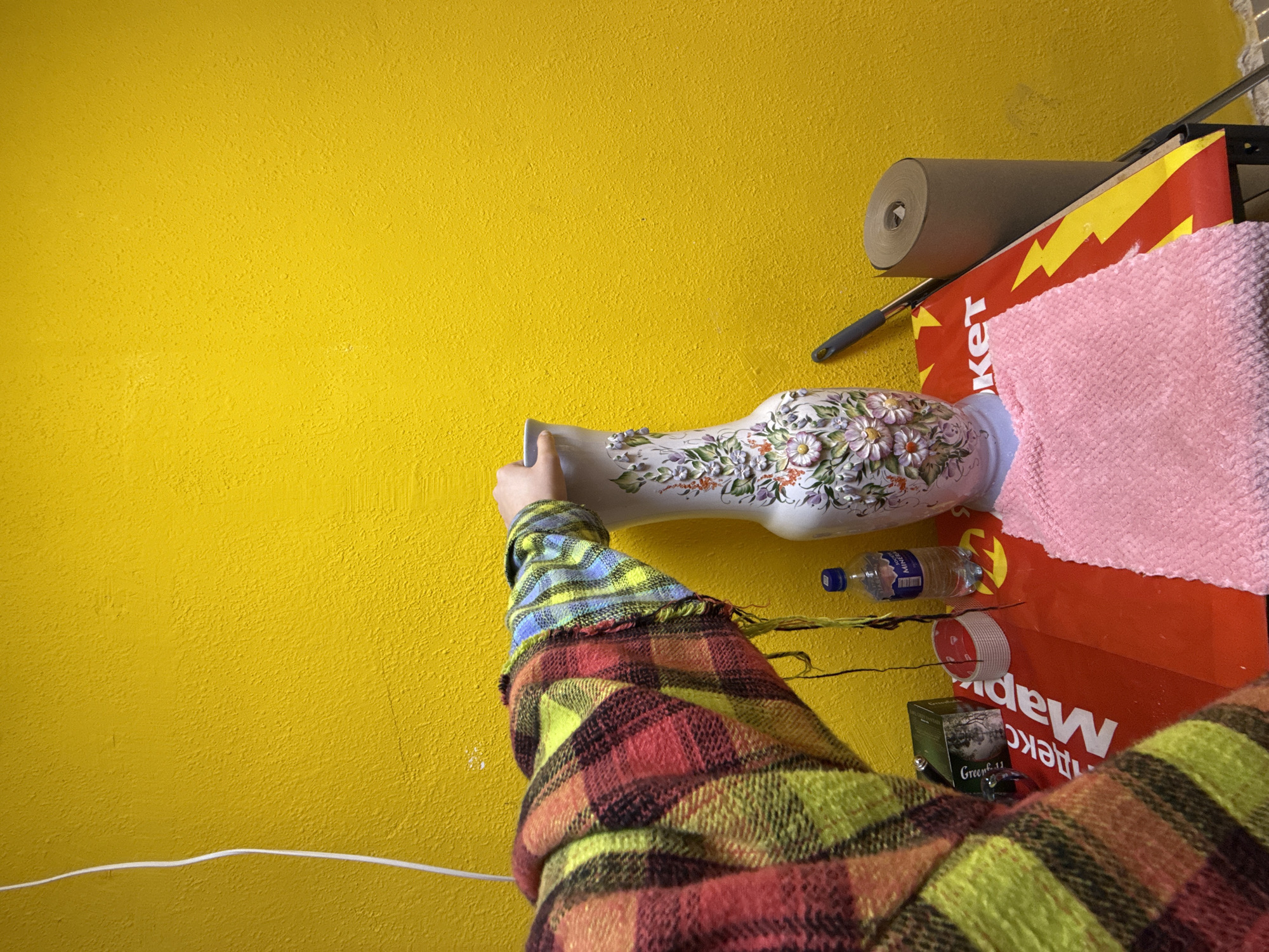 A person wearing a multi-colored plaid shirt is holding a floral-decorated vase against a yellow wall. Nearby, there is a roll of paper, a pink cloth, a water bottle, and a small box labeled "Green.11". A white electrical cord is visible running along the bottom left of the wall. The location is Unknown, Unknown.