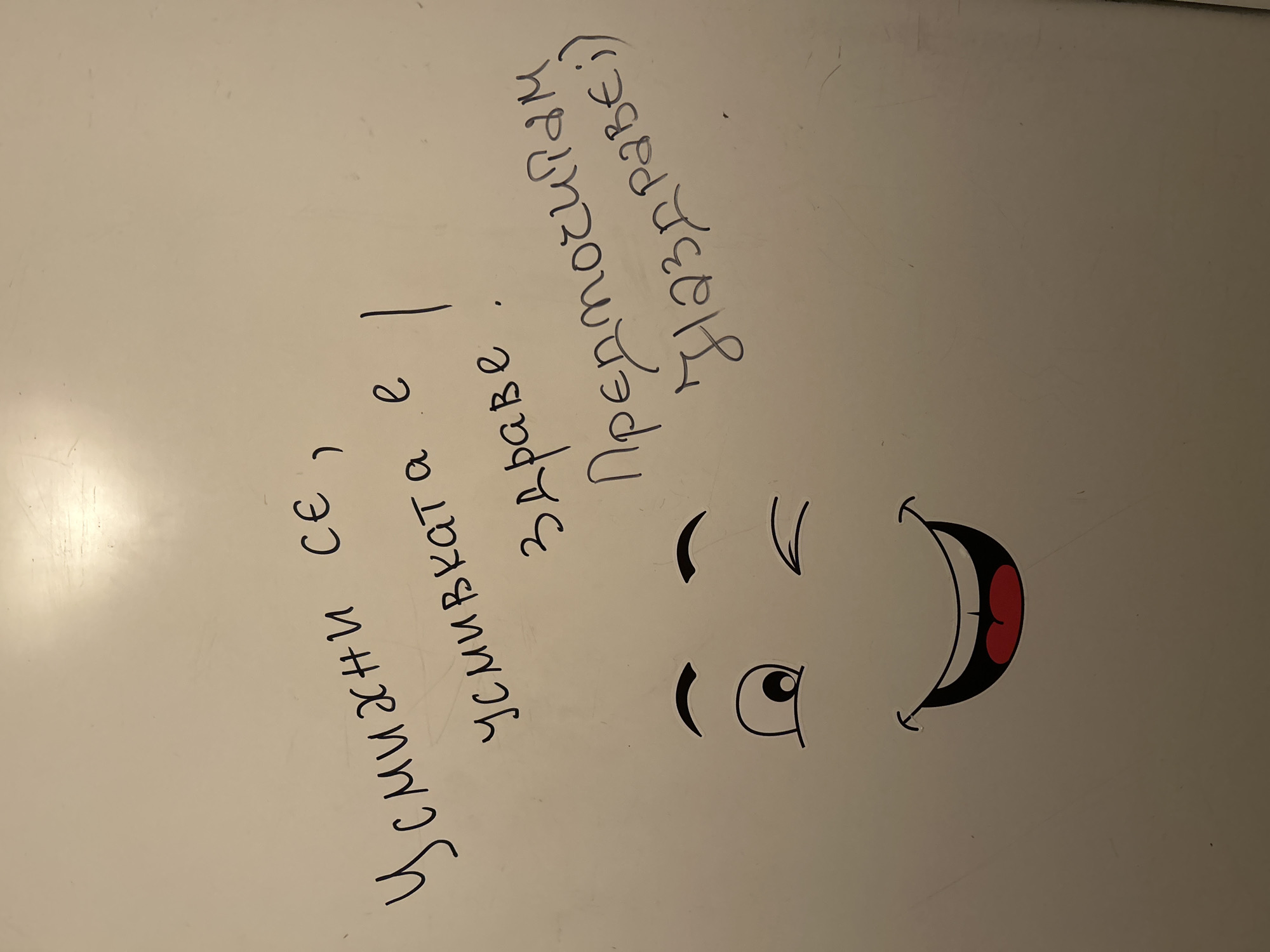 The image shows writing in Bulgarian on a white surface, likely a whiteboard, accompanied by a drawing of a winking face with a big, open mouth. The writing says: "усмихни се, усмивката е здраве. Предпочитам наздраве." This translates to "Smile, a smile is health. I prefer cheers (to good health)". The face is cartoonish and has one eye closed and one eye open, suggesting a wink, with a wide, open mouth. There is no information to place this in Plovdiv, Bulgaria.