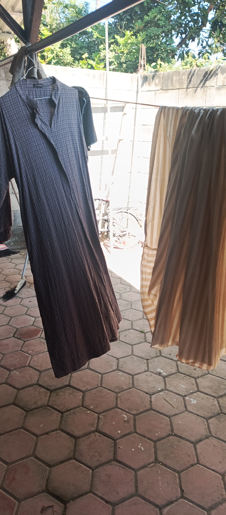 An outdoor laundry drying scene is depicted in Arjasa, Indonesia, with two garments hanging from a clothesline. On the left, a dark, patterned, long-sleeved dress is draped over a hanger. To its right, a lighter-colored, striped fabric is also hanging. The background shows a rough-textured concrete wall, lush green foliage, and a glimpse of a bicycle. The ground is paved with hexagonal tiles in reddish-brown hues, with some debris scattered on the surface. The lighting suggests it is daytime, with shadows cast by the hanging clothes.