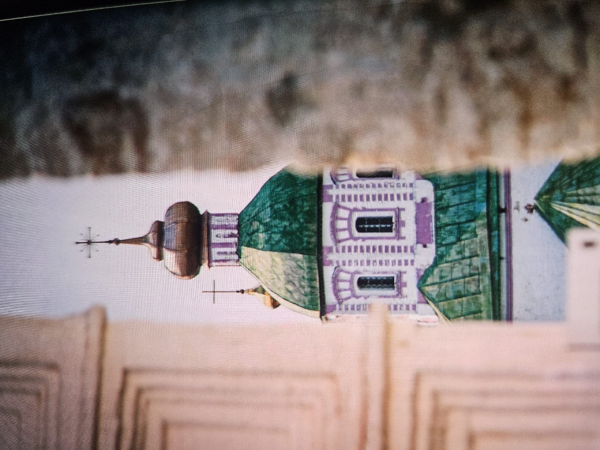 The image shows a close-up of a church dome, likely belonging to the Dormition Cathedral in Vladimir, Russia. The dome is copper-colored and topped with a cross. Below the dome, the church features green roofing and white walls with purple accents and arched windows. The structure is partially obscured by blurred foreground elements, which appear to be architectural details. The overall impression is of a detail captured from a specific vantage point within the city.