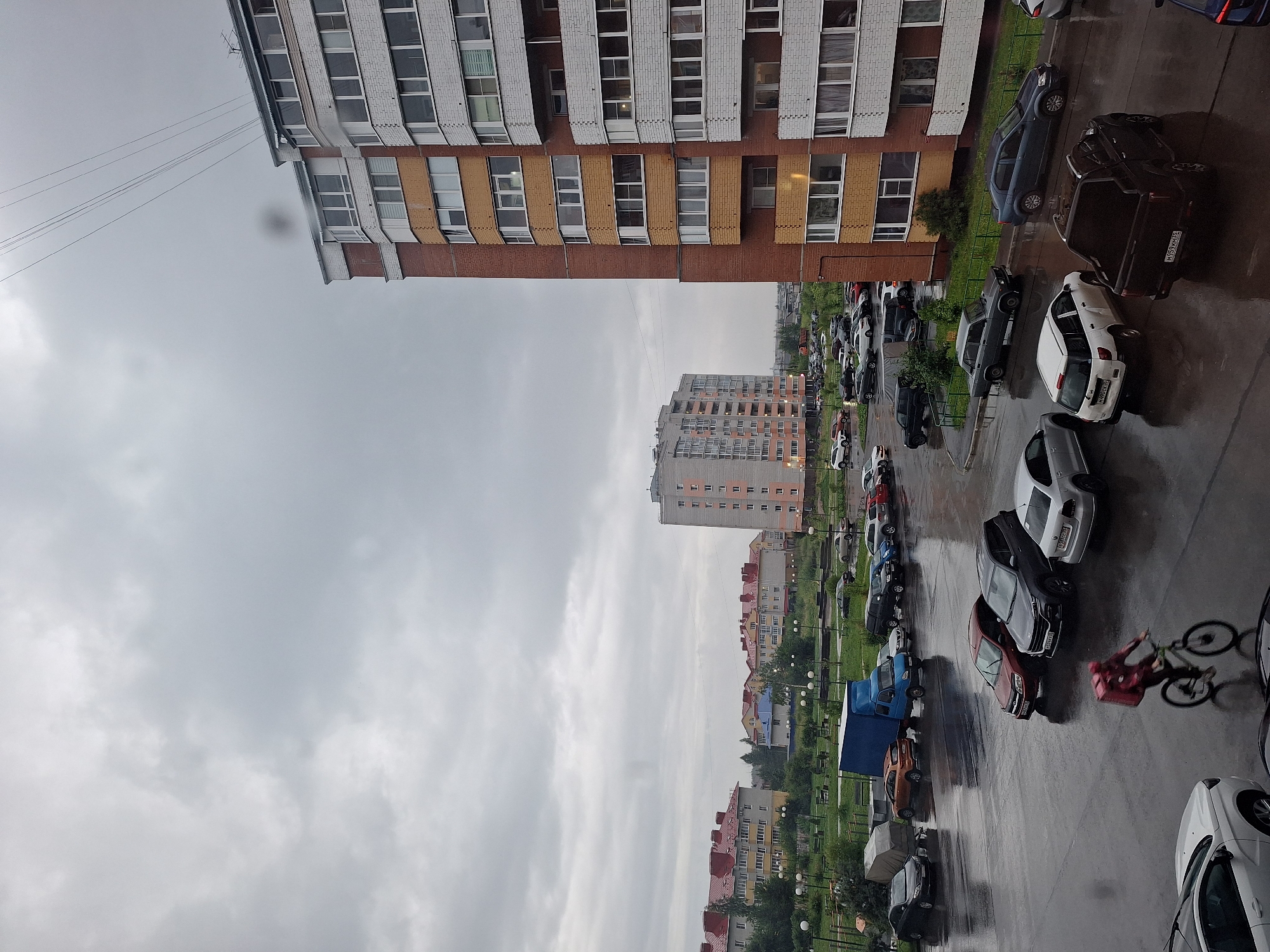 The image captures an overcast day in Vologda, Russia, with a focus on a residential area characterized by multi-story apartment buildings and a wet, paved parking lot. In the foreground, a person on a bicycle, wearing a red jacket and a large pink backpack, cycles across the wet asphalt. Several cars are parked in the lot, including a dark-colored pickup truck with a license plate visible, a white hatchback, a dark grey sedan, and other vehicles in varying shades of grey, red, and blue. A blue truck is parked further back. Beyond the parking lot, a green grassy area with lampposts and benches separates the foreground from a series of apartment buildings. The buildings are a mix of styles and colors, with red and yellow brick facades and numerous windows. In the background, a taller, light-colored apartment building stands out. The sky is heavily clouded, contributing to the muted, diffused light of the scene. The wet surface of the parking lot suggests recent rain.