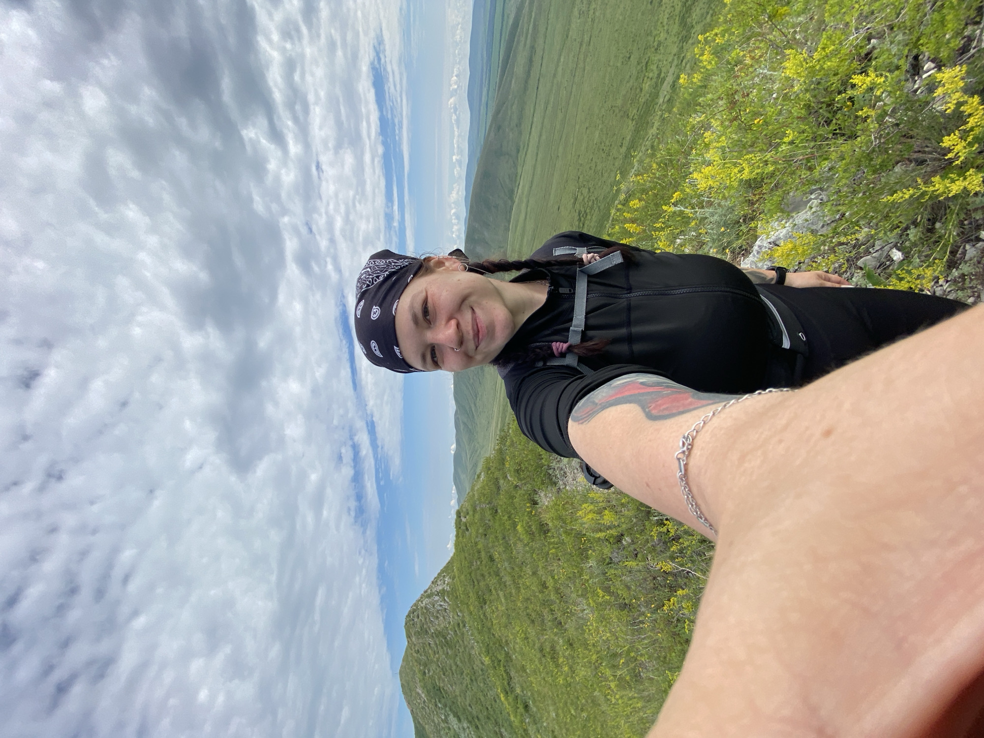 A medium shot captures a young woman with dark hair styled in braids, wearing a black bandana and a black athletic jacket. She is smiling and looking at the camera, holding up her left arm to take a selfie. Her arm shows a tattoo on the upper part and a silver bracelet around her wrist. She is outdoors, on a grassy, hilly landscape with patches of yellow wildflowers. In the background, rolling green hills stretch into the distance under a cloudy sky. The scene appears to be a natural setting, possibly a hiking trail or a mountain ridge. The location is identified as Verkhnyaya Chernorechka, Russia.
