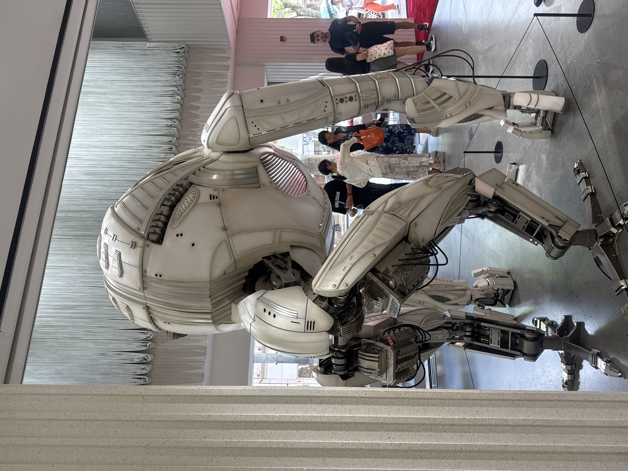 The image shows a large, white, intricate robot sculpture positioned inside a modern, well-lit building. The robot, with a bulbous head and multiple articulated limbs, stands on a dark grey floor. A fluted white column is visible in the foreground on the left. Behind the robot, several people are observing the artwork. In the background, there is a white, textured wall with vertical pleats, and glimpses of the city through windows, suggesting an urban environment. The overall atmosphere appears to be that of an art exhibition or a public space showcasing contemporary art. The location is Shanghai, China, as indicated by the context provided.