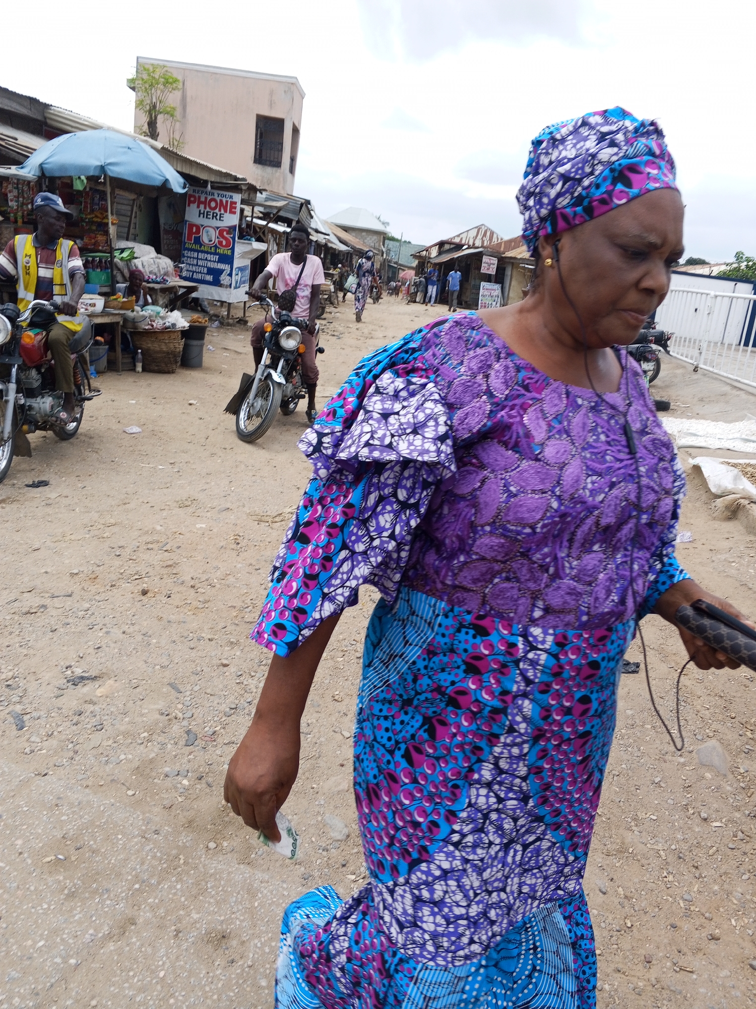 This media file captures a street scene in Zango-ndaji, Nigeria. The primary subject is a woman in the foreground, walking and looking downwards. She is wearing a vibrant, patterned outfit in shades of purple, blue, and pink, with a matching headscarf. She also has earbuds in and is holding a dark-colored object in her right hand. Her left hand is extended downwards, holding a small amount of what appears to be cash.
In the background, other people and objects are visible, suggesting a bustling market or street. Two men are riding motorcycles, one on the left and one in the middle ground. The man on the left wears a yellow and blue vest over a white shirt and a blue cap. The man in the middle ground wears a pink t-shirt. Various market stalls are set up, with one featuring a blue umbrella and goods displayed on tables. A sign with text is visible on one of the stalls.
The ground is dusty and sandy, characteristic of a dry, possibly rural or semi-urban environment. The buildings in the background appear to be simple structures, some with corrugated metal roofs. The sky is overcast, indicating a cloudy day. The overall atmosphere suggests an ordinary day in a busy Nigerian community. The visible text on the sign reads "REPAIR YOUR PHONE HERE" and "POS".