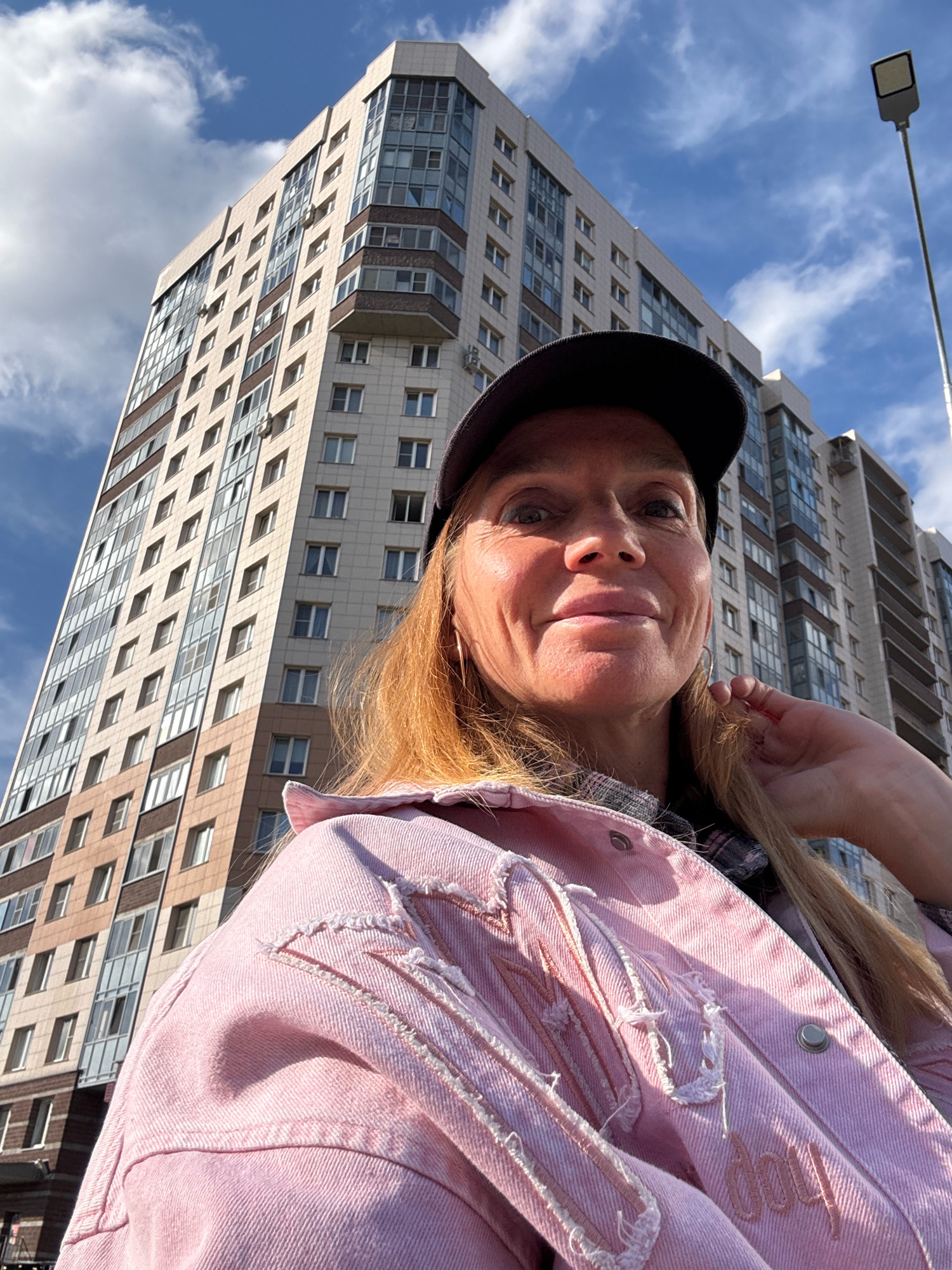 A woman is taking a selfie outdoors in Ladozhskoe Ozero, Russia. She is wearing a pink denim jacket and a black baseball cap. A tall, modern apartment building dominates the background, with a clear blue sky and scattered white clouds above. A street lamp is visible on the right side of the frame. The woman is looking at the camera, with her right hand touching her hair.