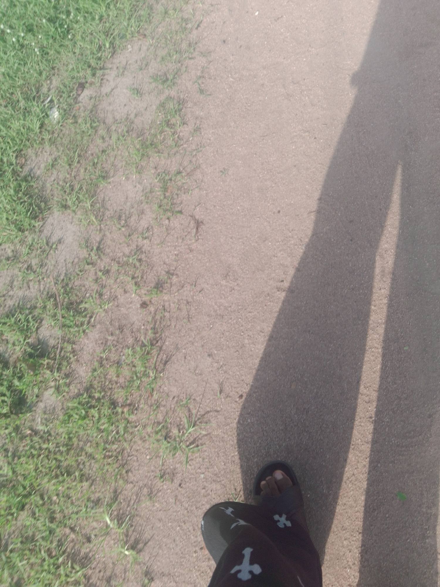 The image shows a person's left foot wearing a dark sandal with a white cross pattern on the upper strap, standing on a paved surface. The foot is casting a long shadow to the right, indicating the sun is to the left. The paved surface appears to be a road or path. To the left of the paved surface, there is an area of sparse green grass and sandy soil. The scene is likely outdoors and during the daytime. The context provided suggests this scene is located in Jalingo, Nigeria.