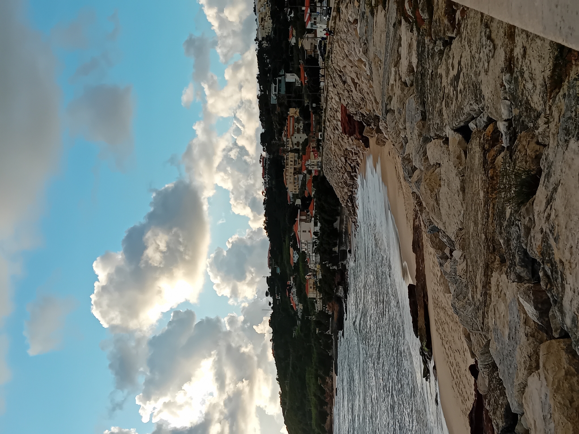 The image captures a coastal scene featuring a rocky embankment on the right side of the frame, extending diagonally downward. To the left of the embankment is a narrow beach with a shoreline where the water meets the sand. Further inland, a hillside dotted with buildings is visible, leading up to a skyline dominated by a partly cloudy sky. The clouds are puffy and white, contrasted against the blue sky. The presence of buildings suggests a residential or urban area along the coast. The overall atmosphere conveys a sense of a coastal community nestled into a hilly landscape under a partly cloudy sky.