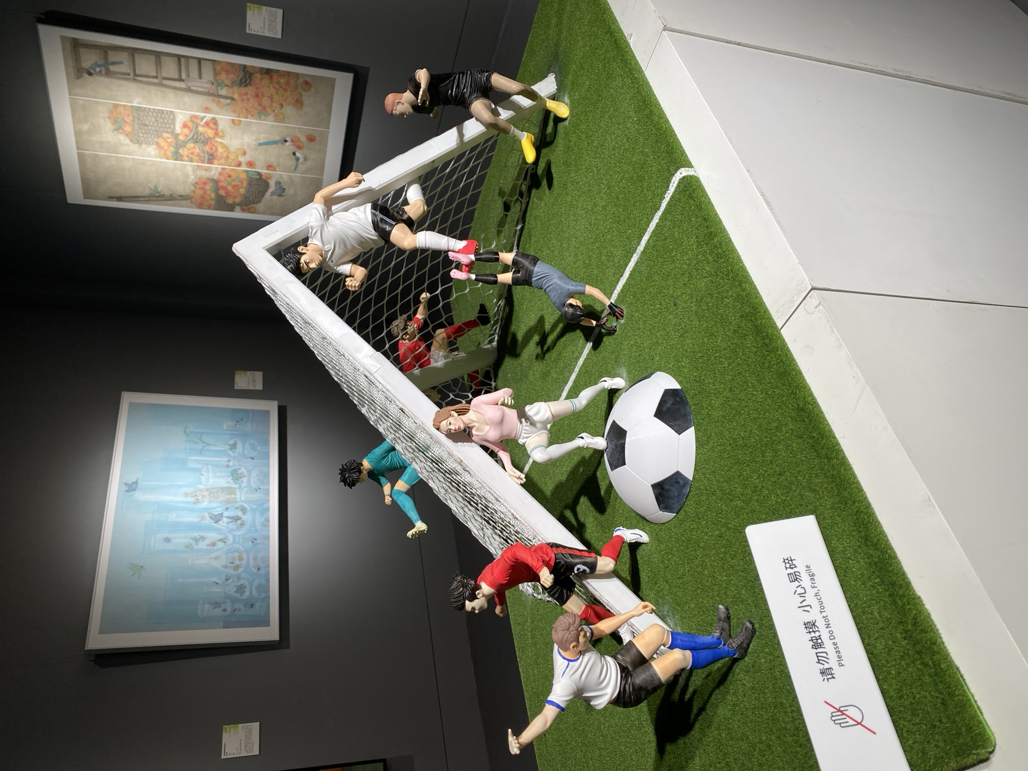 The image displays a diorama of a soccer game, likely in a museum setting in Yuzhong District, China, as indicated by the presence of a "Please Do Not Touch, Fragile" sign in both English and Chinese. The diorama features several miniature figurines of soccer players in dynamic poses on a green, artificial turf field. The scene is centered around a white soccer goal with a net.
The figurines represent players in action: one is jumping towards the goal, another is kicking, one is falling, and others are positioned around the field. They are dressed in various soccer attire. A large, black and white soccer ball is prominently placed on the field. In the background, two framed artworks hang on a dark gray wall. One painting depicts a collection of glass containers with various items inside, under a light blue sky. The other artwork features baskets overflowing with fruit and a ladder. The diorama itself is placed on a white, multi-paneled structure, suggesting a display pedestal.