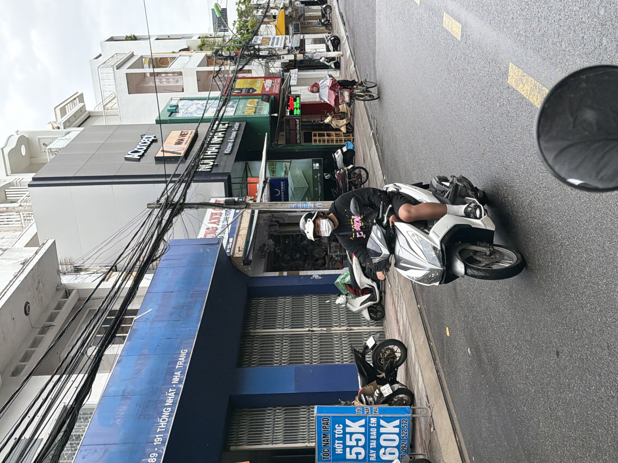 The image depicts a street scene in Nha Trang, Vietnam. A person wearing a white helmet and a face mask is riding a white scooter. They are wearing a dark-colored hoodie with some pink lettering and black shorts, with white socks and patterned slides. Another person is riding a bicycle, wearing a red and pink poncho. Several other scooters and motorcycles are parked along the sidewalk. Buildings line the street, with various signs and storefronts visible, including one that reads "Aconcept" and another with Vietnamese text. The street is wet, suggesting it may have recently rained. Overhead, a tangled mess of electrical wires is prominent against the overcast sky. A blue sign with Vietnamese text and prices is also visible on the left side of the image.