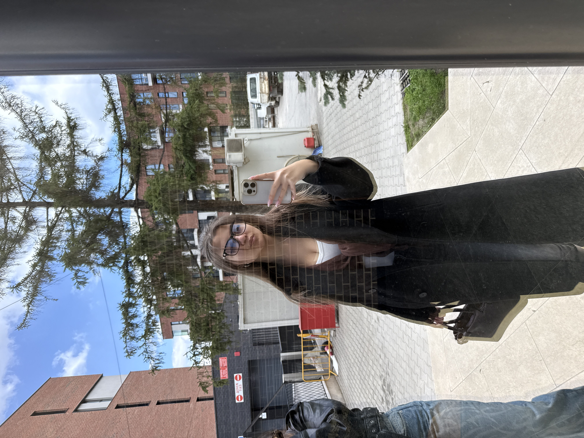 A photograph shows a young woman reflected in a window, taking a selfie with her smartphone. She is wearing glasses, a white top, and a black coat. Below her reflection are a dark leather jacket and a pair of blue jeans. The background features a cityscape, including brick buildings, trees, paving stones, a white van, and various signs. The location appears to be Sofia, Bulgaria.