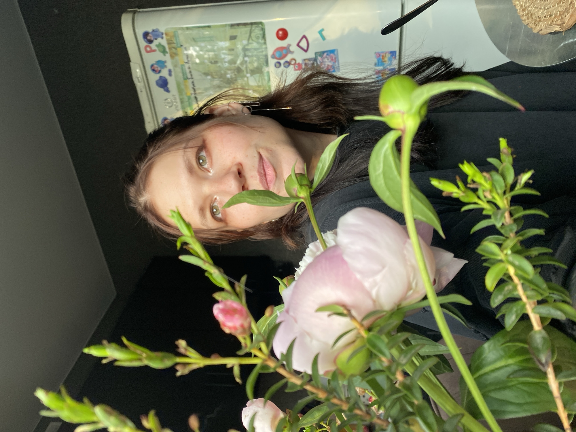 A young adult female is depicted in a medium shot indoors. She has long dark hair, light-colored eyes, and is wearing a black long-sleeved top and two silver-toned chain earrings. Her gaze is directed slightly upwards and to her left. A bouquet of flowers and green foliage, including at least one light pink peony and multiple buds, is positioned in the immediate foreground, partially obscuring the woman. The background features a dark, unidentifiable surface on the left and a white refrigerator on the right. The refrigerator door is adorned with multiple colorful magnets and stickers. Reflections on the refrigerator display an outdoor scene with foliage and structural elements, possibly through a window. A metallic spoon and a woven coaster are visible on a surface in the lower right corner. The scene is illuminated by natural light, casting highlights on the woman's face and the refrigerator.