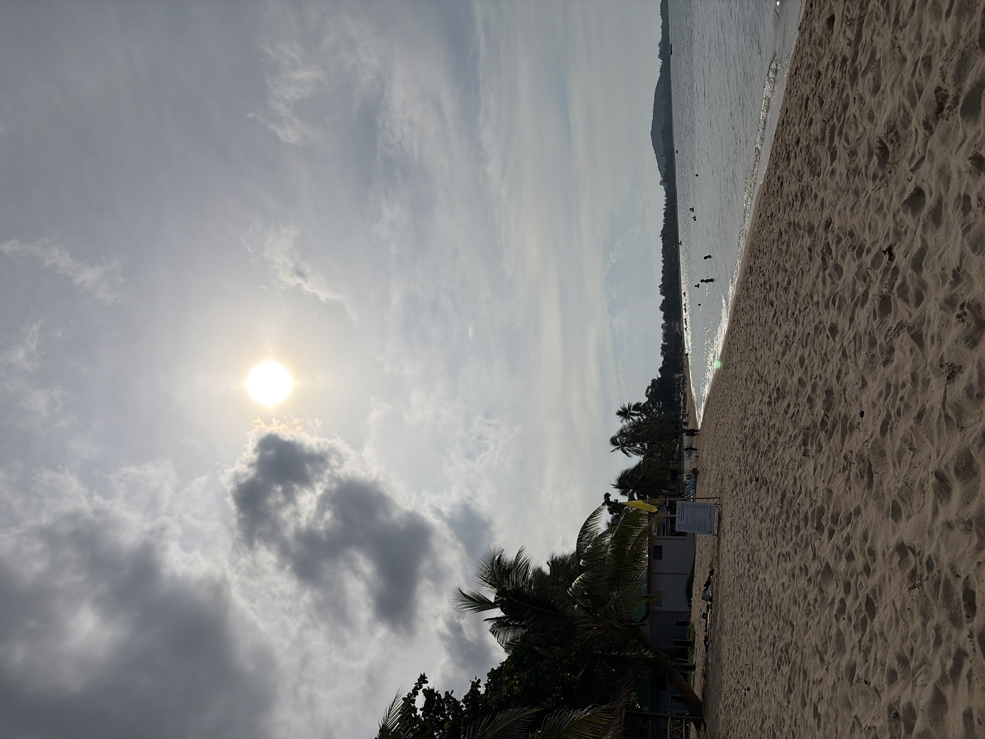 The image depicts a sandy beach on the island of Ko Samui, Thailand. The sun is visible in the sky, partially obscured by clouds, casting a bright light. Palm trees line the edge of the beach, with a few buildings and a yellow flag visible among them. Several people are present on the beach and in the shallow water, some swimming and others walking along the shoreline. The sea is calm, and a distant shoreline with hills can be seen. The sand in the foreground is textured with ripples, suggesting it is a well-used beach.