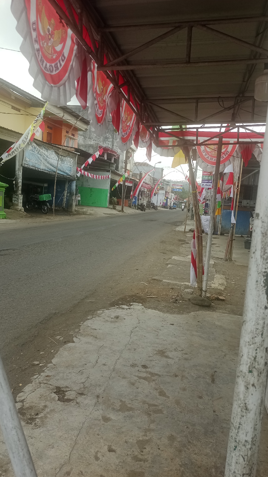 The image captures a street scene in Cikalong, Indonesia, characterized by a prominent display of Indonesian flags and celebratory banners. These decorations, predominantly red and white, hang from structures lining the street, suggesting a national holiday or event. The scene includes a paved road with parked motorcycles and a few distant vehicles. Buildings with a mix of architectural styles, including what appears to be a shop or workshop, are visible along the roadside. The overall environment is that of a moderately built-up urban or semi-urban area.