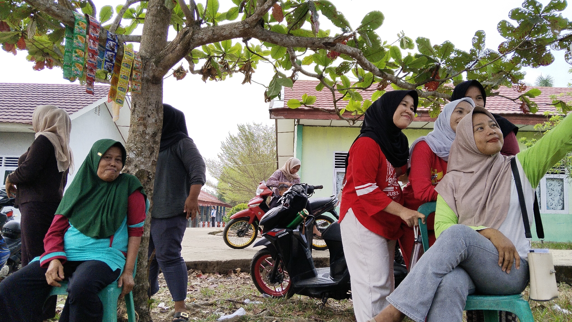 A group of women are gathered outdoors, likely in Margatunggal, Indonesia, near a building with a light green facade and red-tiled roof. A large tree with green leaves and some red fruits dominates the foreground, with several packets of snacks hanging from its branches. In the center, a woman wearing a green hijab and a teal and red top sits on a green chair, looking towards the camera with a relaxed expression. To her left, a woman in a brown hijab stands. To her right, a woman in a black hijab stands near the tree trunk. Further back, several other women are visible, some near motorcycles. One woman, wearing a red top and white pants, is taking a selfie with two other women in the background. The ground is covered with fallen leaves and debris, and a paved pathway is visible. Two motorcycles, one red and one black, are parked in the scene.
