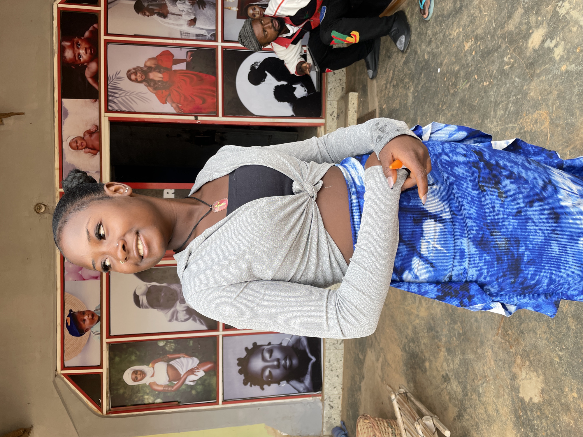 A young woman stands in front of a wall adorned with various photographs, likely a photography studio, in Barakin Akawo, Nigeria. She is wearing a gray, long-sleeved, knotted top, a black bra, and a blue patterned skirt or wrap. She has dark hair pulled back in a bun and is smiling slightly. She is holding an orange object in her hands. Behind her, there are several framed photographs of babies, couples, and individuals, indicating the professional nature of the establishment. In the background, a man is visible sitting on a step. The ground is concrete.