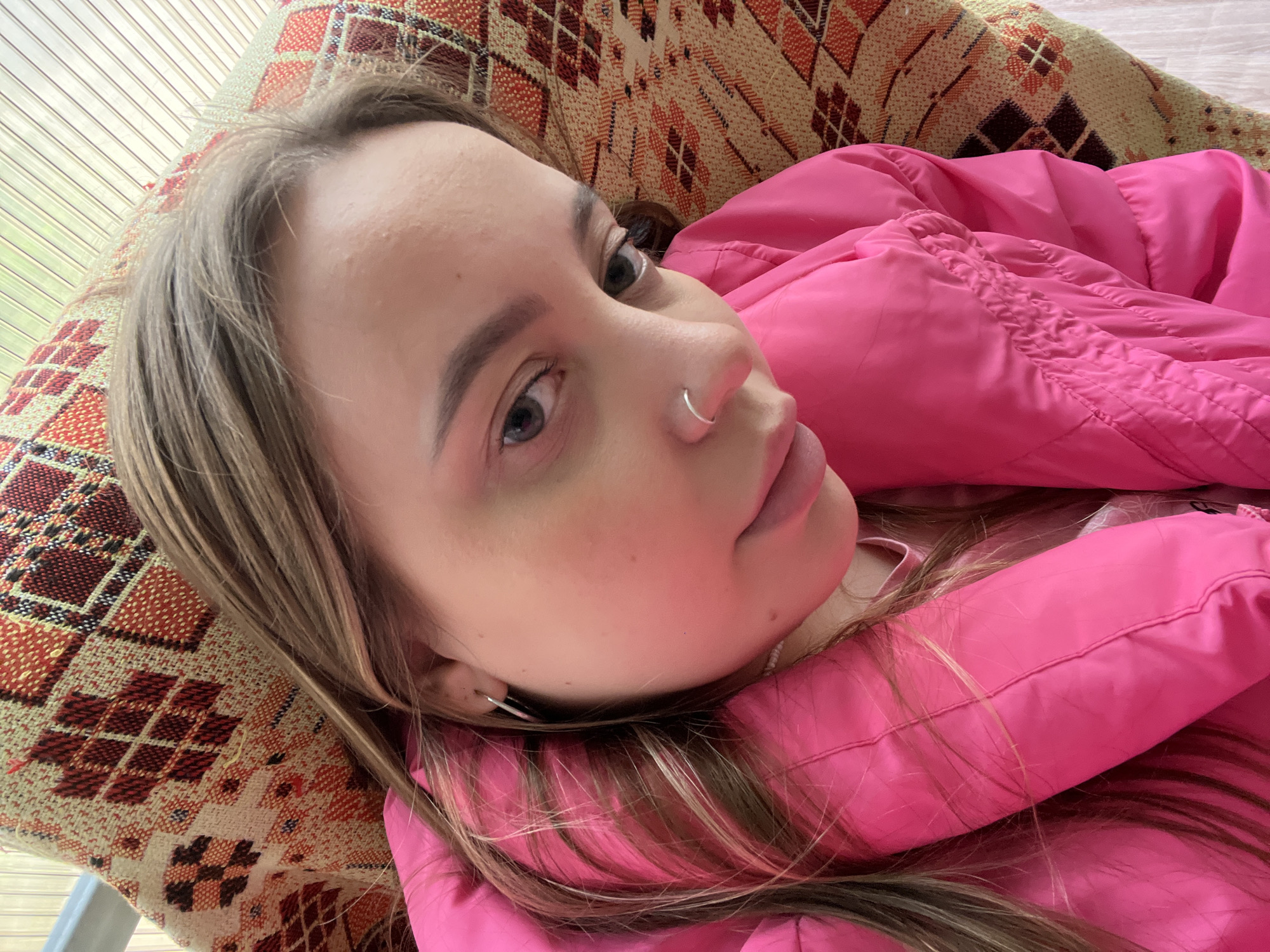 A young woman with fair skin, light brown hair, and gray eyes is lying on a patterned fabric surface. She is wearing a bright pink padded jacket. She has a silver nose ring and a small hoop earring. The background includes a light-colored paneled wall or window. The location is not evident in the image.