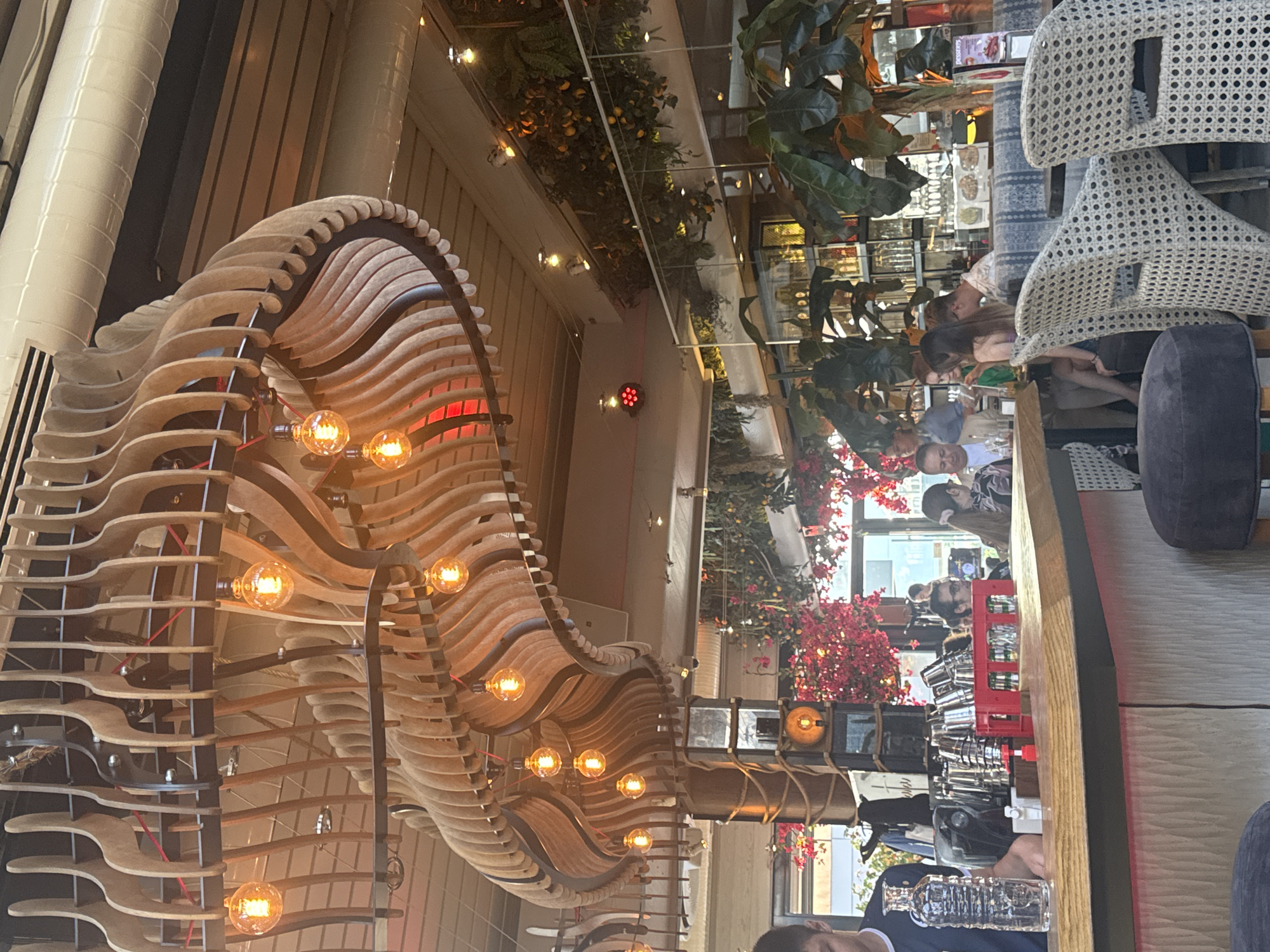 This image captures the interior of a restaurant or bar located in Varna, Bulgaria. The scene is dominated by a large, ornate chandelier constructed from numerous light-colored wooden slats arranged in a wave-like, curvilinear pattern across the ceiling. Multiple spherical, exposed-filament light bulbs, emitting a warm orange glow, hang from the chandelier, some connected by visible red wiring. The ceiling above consists of light-colored panels, featuring white ventilation ducts and recessed spotlights.
In the mid-ground, substantial arrangements of lush greenery, including plants bearing small, yellow, fruit-like objects, line overhead structures and walls. Prominent clusters of vibrant pink flowering plants are visible towards the left and right center. Sections of clear glass partitioning are present, separating different areas.
Several patrons are seated throughout the establishment. In the right mid-ground, individuals occupy tables with light-colored, woven chairs; one person with dark hair tied in a ponytail is facing away. Another individual wearing glasses is visible deeper within the background.
The foreground features a curving wooden bar counter with a textured, vertically striped light-colored front. On the far left, a person, partially visible and wearing a dark shirt with a white collar, stands behind the bar. A clear glass bottle is on the bar surface. Behind the bar, various items include stacked metal cups or shakers, a red plastic crate holding small glass bottles, and a tall, dark column wrapped with rope, featuring a central illuminated circular element. Dark, upholstered bar stools are partially visible in the immediate foreground.