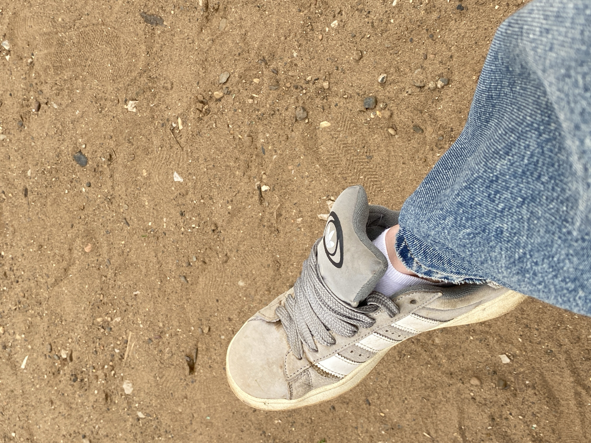 A close-up, overhead shot shows a person's left foot wearing a grey Adidas sneaker with white stripes, a white sock, and blue jeans. The sneaker is positioned on a patch of sandy ground, which appears to be part of a park or open area. There are small pebbles and debris scattered across the sand. The image does not provide any specific visual cues to identify the location as Votkinsk, Russia.