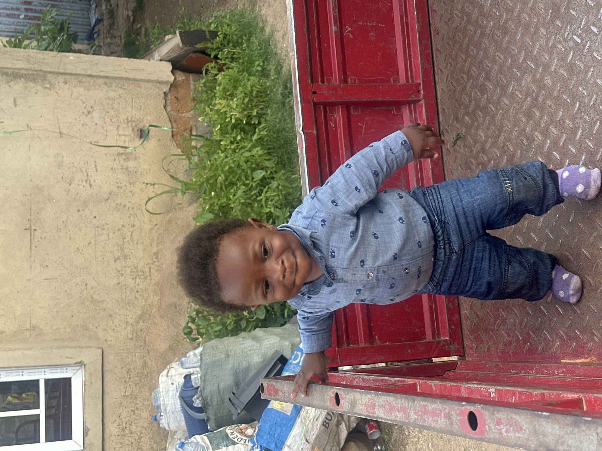 A young Black child, appearing to be about two years old, stands in the bed of a red truck. The child is wearing a light blue collared shirt with small elephant patterns and dark blue jeans. The child's feet are clad in light purple socks with white polka dots. The child is holding onto the red metal side of the truck bed with their right hand, which is resting on a protruding bar. The truck bed has a diamond plate metal floor and red metal sides. In the background, a weathered, beige-colored building with a white-trimmed window is visible. To the left of the child, several large bags, some appearing to be made of woven material and others of plastic, are piled up, with some water bottles visible amongst them. Green vegetation grows behind the truck bed and to the right of the building. The scene appears to be outdoors, potentially in a yard or alleyway, consistent with a location in Gura Top, Nigeria. The lighting suggests it is daytime.