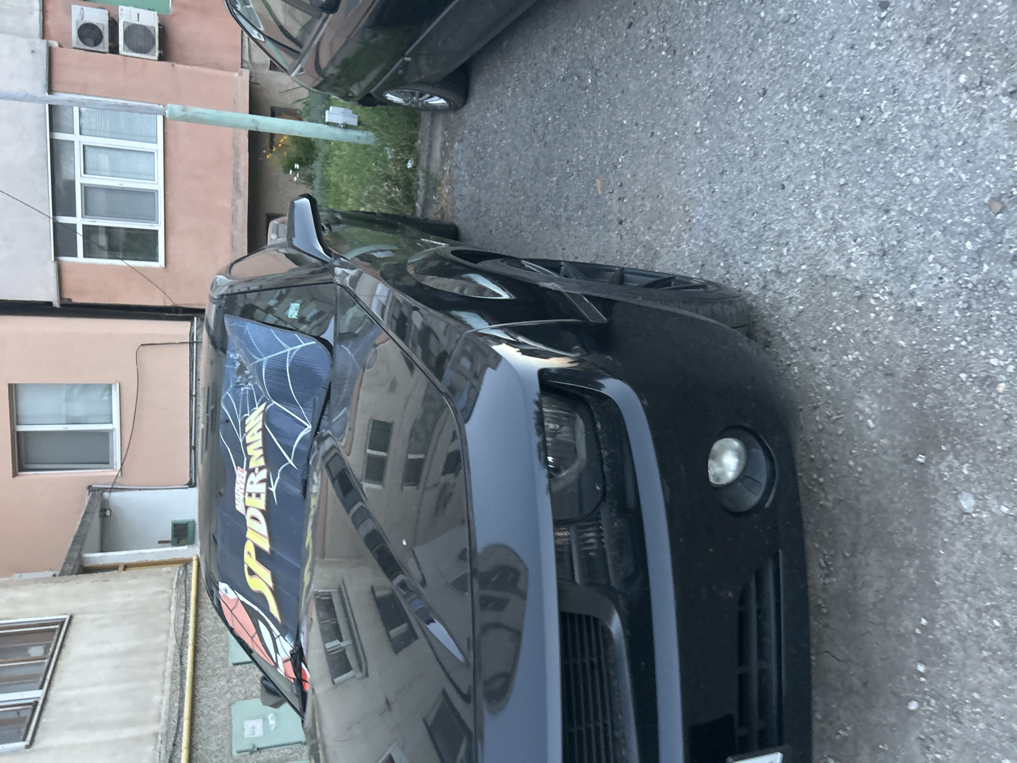 A black Chevrolet Camaro is parked next to a building. The car has a "Spider-Man" decal on the windshield. The building appears to be a residential building with several windows and air conditioning units. The area around the car is paved with gravel.