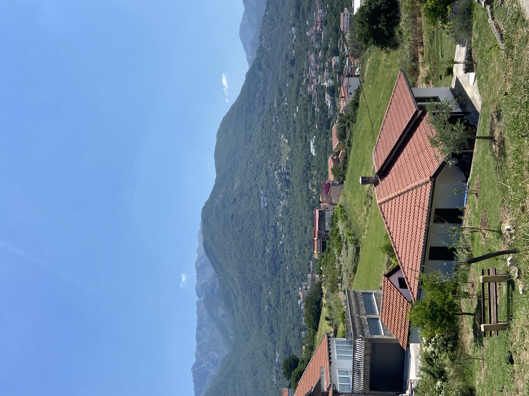 This is a vertical, high-angle outdoor shot of a hillside settlement in Šušanj, Montenegro, on a clear, sunny day. The scene is dominated by the verdant slopes of a large mountain range in the background, dotted with numerous houses and buildings that cascade down the incline. The foreground features several houses with terracotta tiled roofs, set amidst well-maintained green lawns and some trees. One house in the foreground has an olive tree and a wooden bench in its yard. Further down the slope, more houses are visible, some with modern architectural elements. The clear blue sky suggests it is daytime, likely mid-morning or afternoon, with no significant cloud cover except for a wispy white cloud near the mountain. The overall impression is one of a peaceful, residential area nestled into a picturesque natural landscape. There is no visible text in the image.