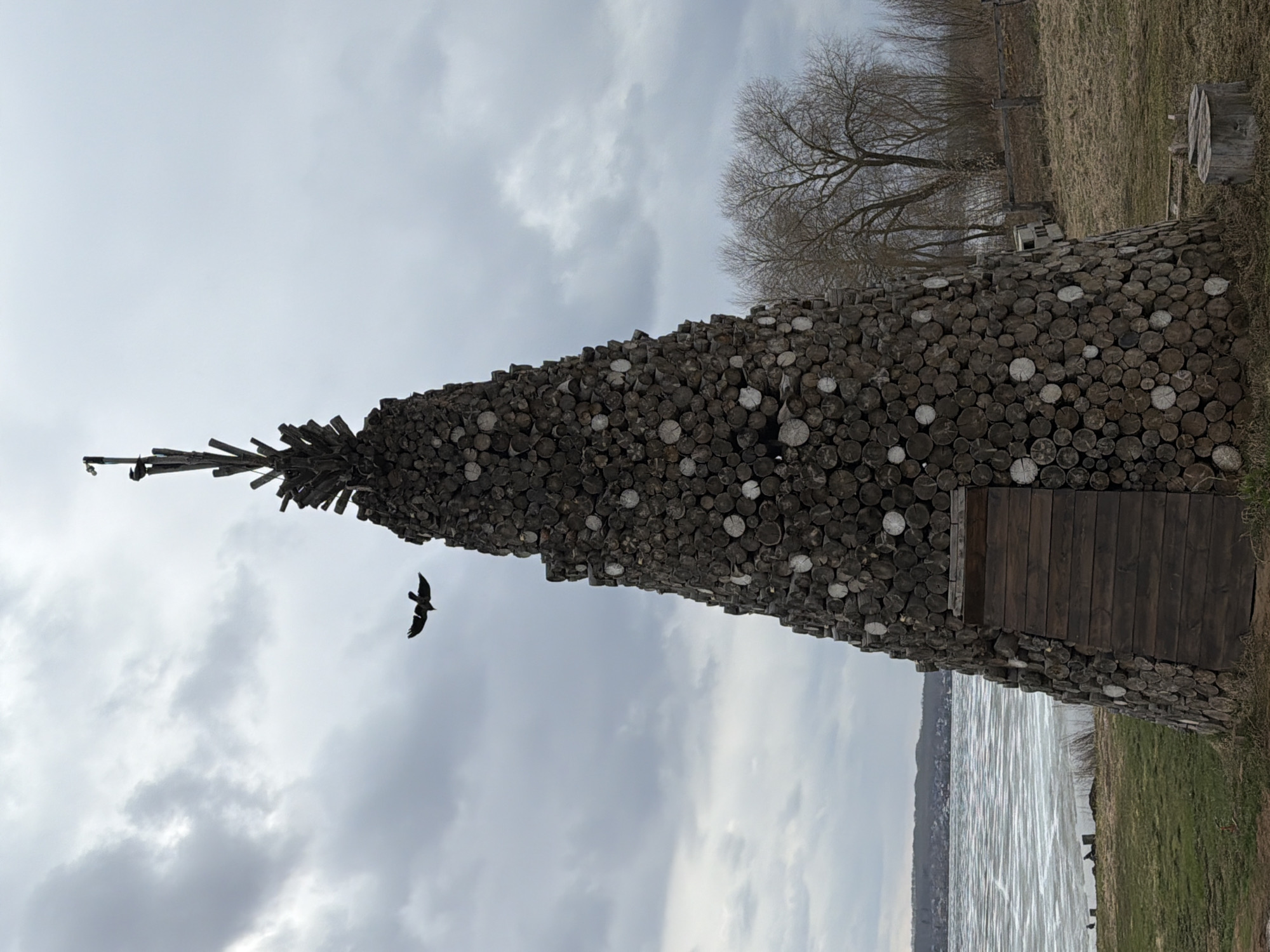 The image depicts a tall, cone-shaped structure constructed from stacked logs, resembling a teepee or wigwam, located near a body of water. The structure appears to have a wooden door. A bird is flying near the top of the structure. The surrounding environment includes grass, trees, and an overcast sky. The location is Votkinsk, Russia.