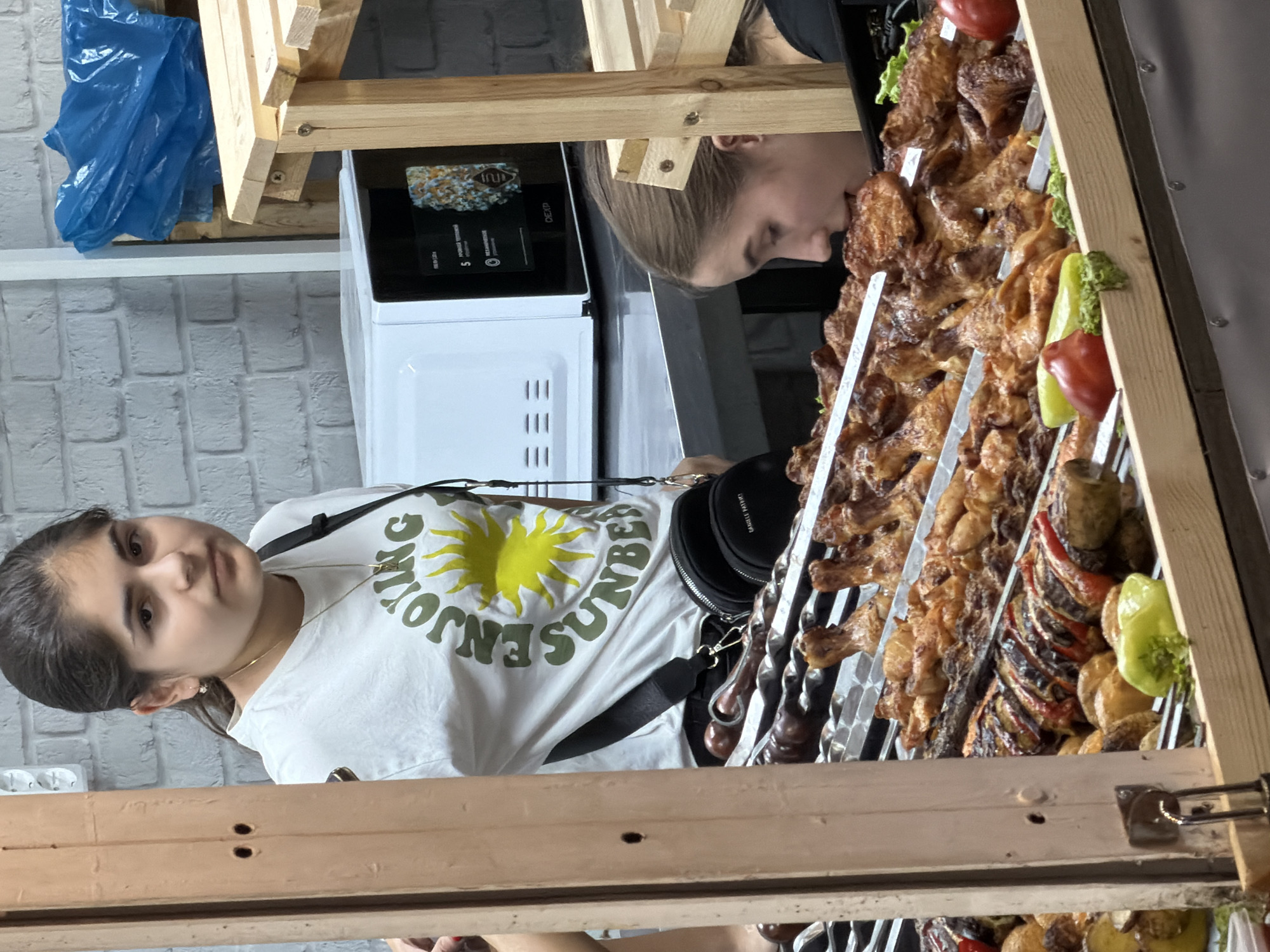 A young woman with dark hair tied back, wearing a white t-shirt with a graphic of a yellow sun and green text, stands behind a display of grilled food. She is looking towards the camera with a neutral expression. In the foreground, skewers of grilled chicken legs and other meats are arranged on a metal rack. Next to the grilled food, there are red tomatoes and green bell peppers. Behind the woman, a white microwave is visible, and to the right, a wooden shelf unit is present. Another young woman is partially visible to the right, leaning in towards the grilled food. The background features a white brick wall. The scene appears to be an outdoor food stall or market.