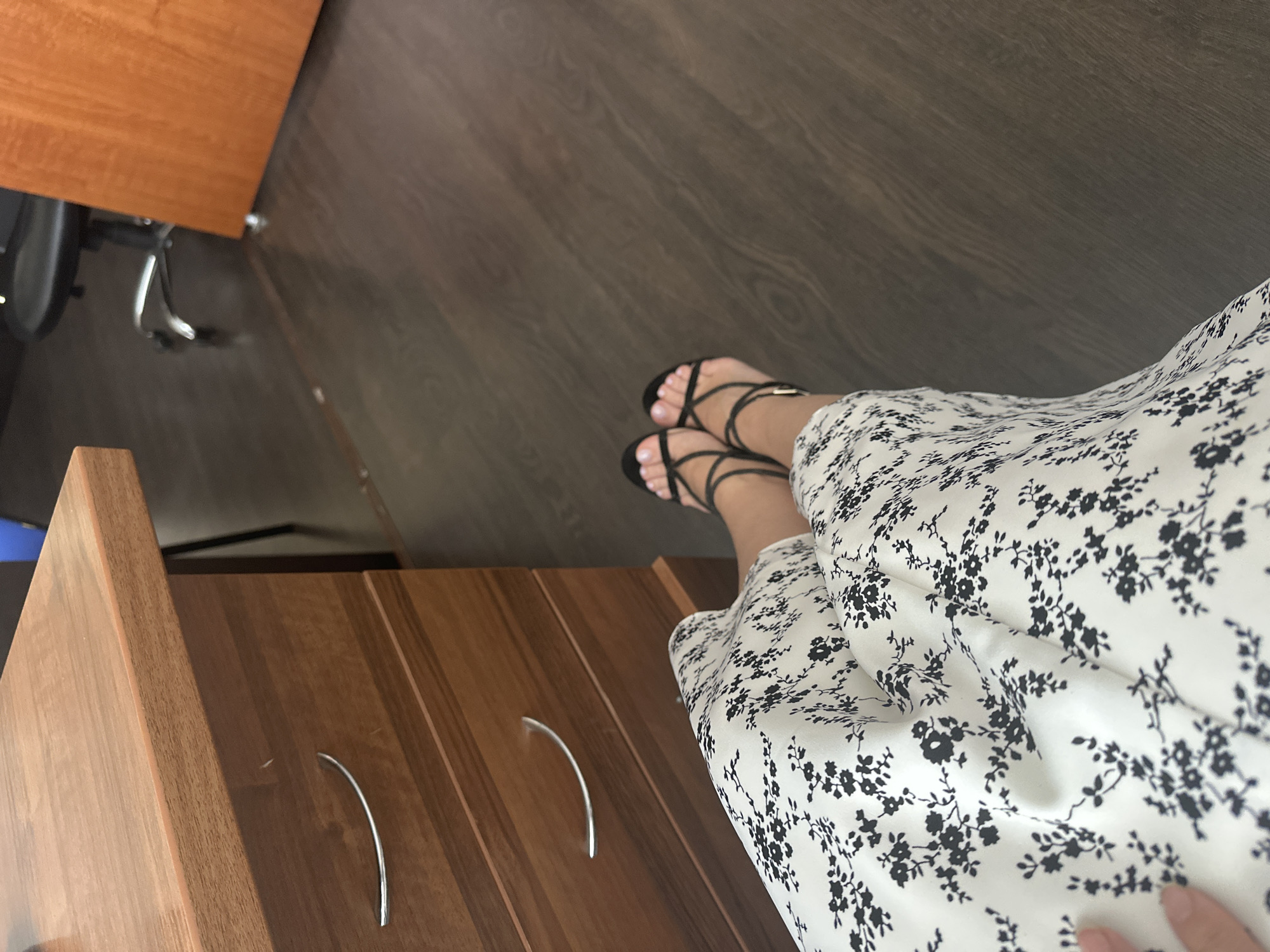 The image captures a close-up view of a person's legs and feet adorned with black strappy sandals, resting on a dark wooden floor. The person is wearing a white dress with a black floral pattern. To the left, a portion of a wooden desk with silver drawer pulls is visible. The background consists of a blurred office environment, with a hint of an office chair and a wooden door. The location is described as Sochi, Russia, though no specific landmarks or context within Sochi are evident. The scene appears to be an indoor office setting, with no other individuals present.