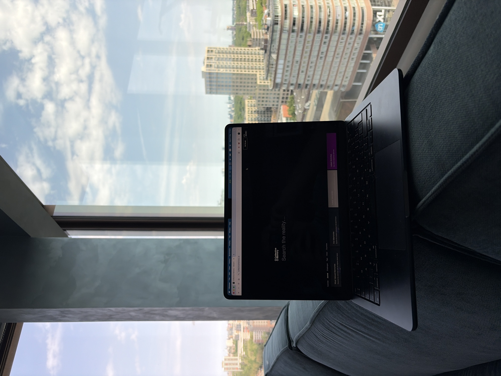 A laptop sits on a blue-gray couch in front of a window in a high-rise building. The laptop screen displays a Chrome browser page. Outside the window, the city of Rijswijk, Netherlands, is visible, with several high-rise apartment buildings under a partly cloudy sky.