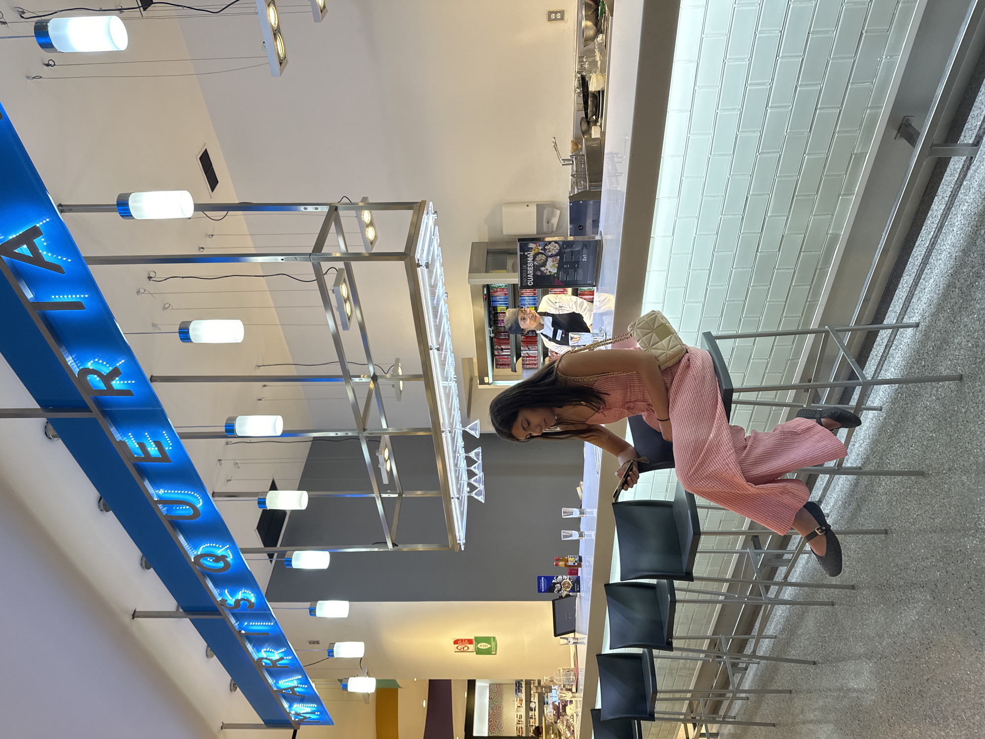 A sideways-oriented full-shot shows a woman seated at a tall bar in what appears to be a food court in Puebla, Mexico.
The woman, with dark hair, is seated on a high stool wearing a light pink, textured, jumpsuit and black shoes with straps. She is leaning toward the bar counter, looking at her phone, which she holds in her hand. Her purse is placed beside her on the bar chair.
The bar area has a long, white countertop with an array of glassware, including wine glasses and tumblers. Behind the bar is a worker, a male wearing a white uniform, facing left toward the viewer. In front of him is a cooler stocked with beverages and what appears to be a menu board.
The environment is characterized by modern architectural elements. Above, there is a blue illuminated sign with words that are not fully readable. A metal frame with illuminated lights extends from the ceiling. Rows of similar high stools line the bar counter, all of which are black. The floor is light gray.
The location is identifiable as Puebla, Mexico, based on the store signage in the background of the image.