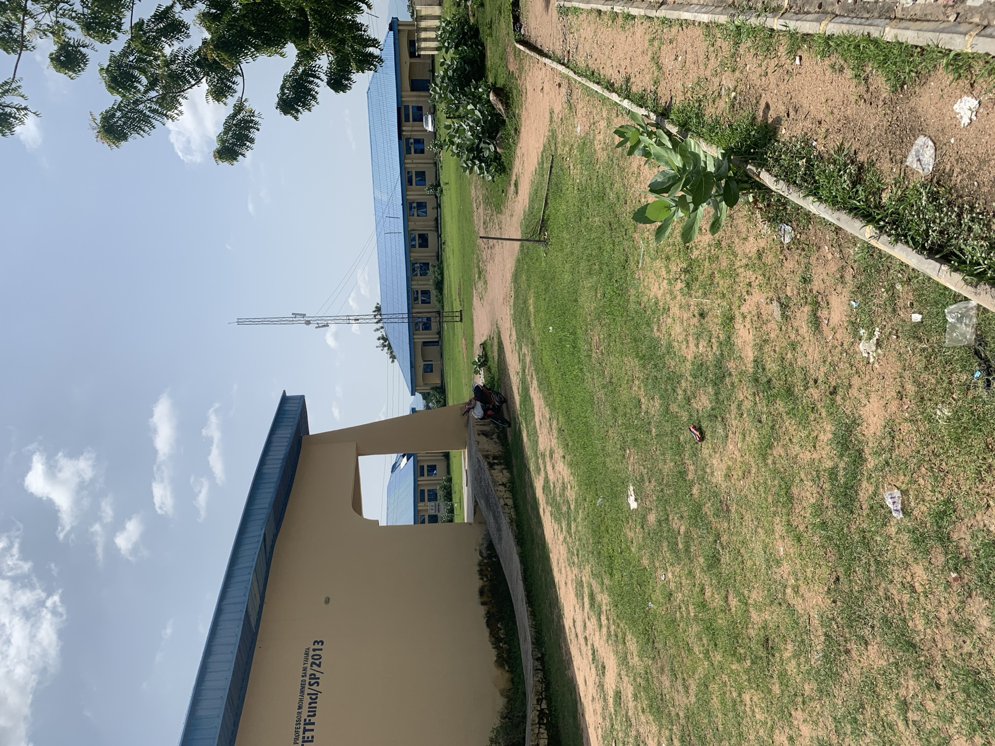 In Jalingo, Nigeria, a sunny day illuminates a section of a university campus. The image captures a light beige building with a blue metal roof, its side featuring signage that reads "PROFESSOR MOHAMMED SANI YAHAYA" and "ETFUnd/SP/2013". A concrete ramp leads up to an open entryway in the building. To the right of the ramp, a red and white motorcycle is parked.
Beyond this building, more campus structures are visible. A long, yellow building with a blue metal roof stretches across the mid-ground, its facade punctuated by numerous windows. A white car is parked in front of this building, and a cellular tower stands tall behind it. The ground is a mix of dry, sandy soil and patchy green grass, with scattered pieces of litter. Lush green trees with dark leaves frame the right side of the scene, their branches reaching towards a sky with scattered white clouds. The overall environment suggests an outdoor, institutional setting on a clear day.