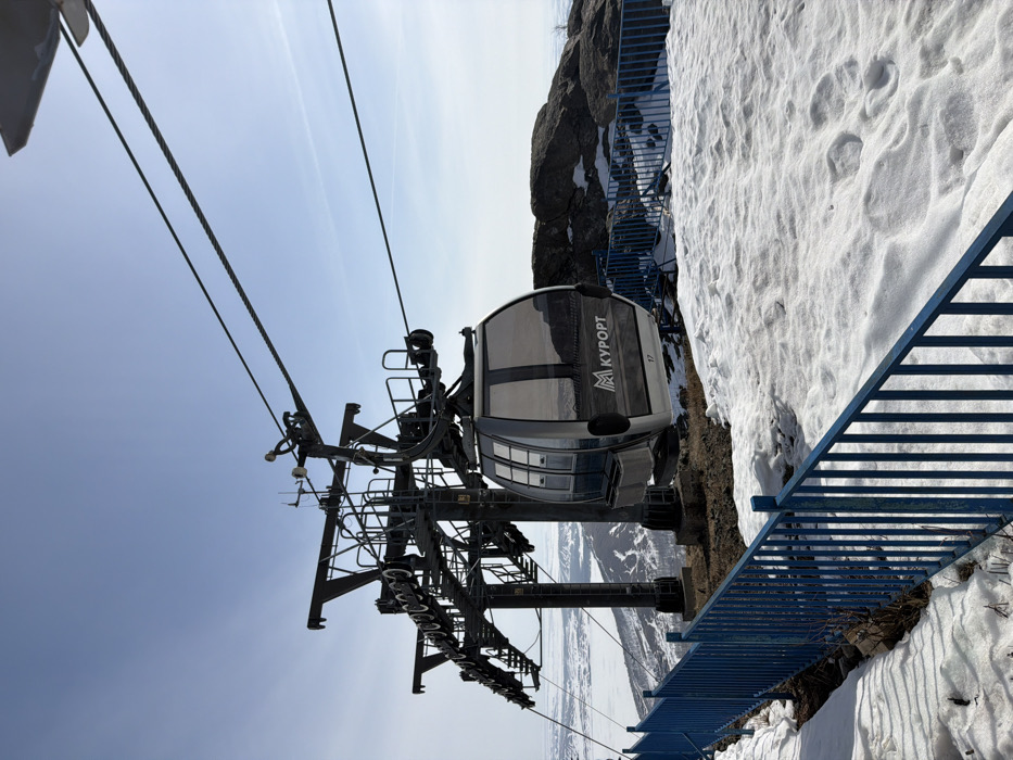 The image displays a ski lift gondola system located in a mountainous, snow-covered environment, identified as Zelenaya Polyana, Russia. A large, dark-grey metal pylon, part of the ski lift structure, stands prominently in the mid-ground, supported by a concrete base. Multiple heavy-duty steel cables extend from the pylon, supporting a modern, silver-grey and black gondola cabin. The cabin is numbered "17" and features the Cyrillic text "КУРОРТ" (Resort) along with a stylized mountain logo on its side, beneath tinted windows.
In the immediate foreground, a blue metal safety fence, composed of vertical railings, runs horizontally across the lower portion of the frame. The ground directly beneath and around the fence is covered with patches of snow, interspersed with areas of exposed dark earth and rock, suggesting melting conditions. Further to the right of the gondola, large, dark rock outcrops are visible.
The background presents a vast panoramic view of a distant landscape, characterized by snow-covered hills and mountains with sparse, bare vegetation. A large, possibly frozen, body of water is visible in the far left distance. The sky above is clear and light blue, indicating fair weather conditions. No individuals are present in the scene.