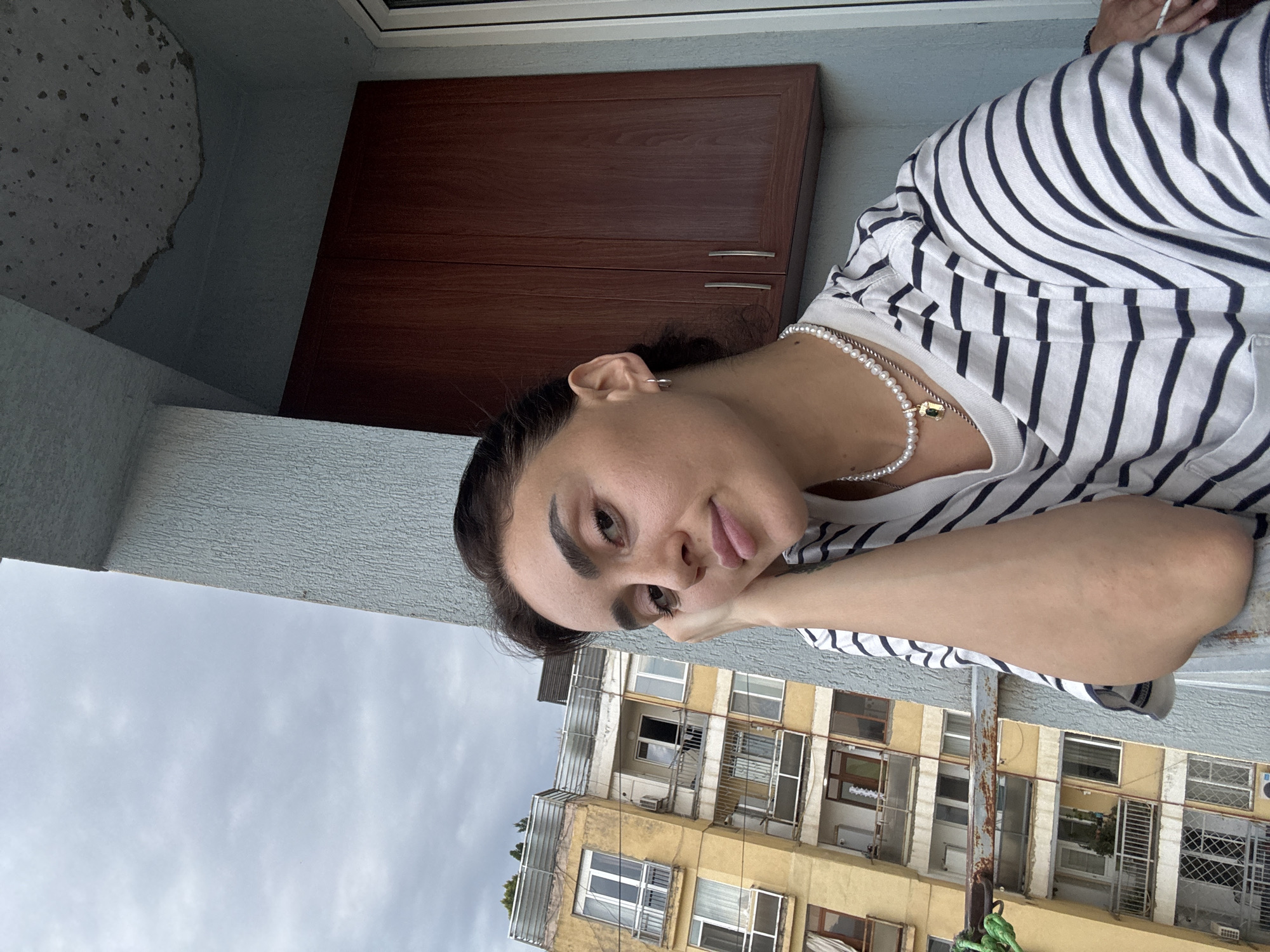 An outdoor, medium shot shows a young woman leaning her head on her hand, looking towards the camera. She has dark hair pulled back and is wearing a striped t-shirt and a pearl necklace with a pendant. Behind her, a yellow apartment building with balconies and windows is visible. The sky is overcast. The scene appears to be on a balcony, judging by the railing and the adjacent wall with cabinets. The architectural style of the building suggests it could be located in the city of Tbilisi, Georgia.