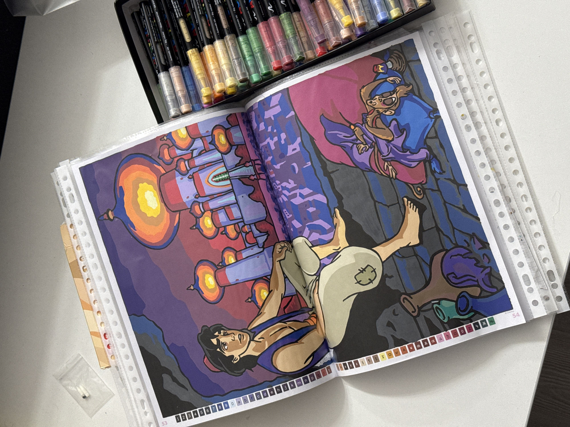 The image shows an open coloring book with a vibrant illustration of characters from Disney's Aladdin. In the foreground, Aladdin is depicted lying down, looking upwards with a concerned expression. Above him, a monkey character, likely Abu, is shown in a blue robe, seemingly holding onto Aladdin. The scene features a fantastical setting with a large, illuminated structure resembling a palace or temple in the background, rendered in warm oranges, yellows, and purples. The overall style of the illustration is cartoonish and colorful, characteristic of animation.
A set of markers is visible at the top of the image, placed in a black box. The coloring book pages are protected in plastic sleeves, with the numbers "53" and "54" visible at the bottom edge of the pages, indicating page numbers. A small plastic bag containing two small white objects is placed on the table next to the coloring book. The image appears to be taken indoors, on a white surface, likely a desk or table, under artificial lighting. The presence of the Disney logo suggests this is an official coloring book. The scene evokes a sense of adventure and fantasy, possibly a moment from the movie.