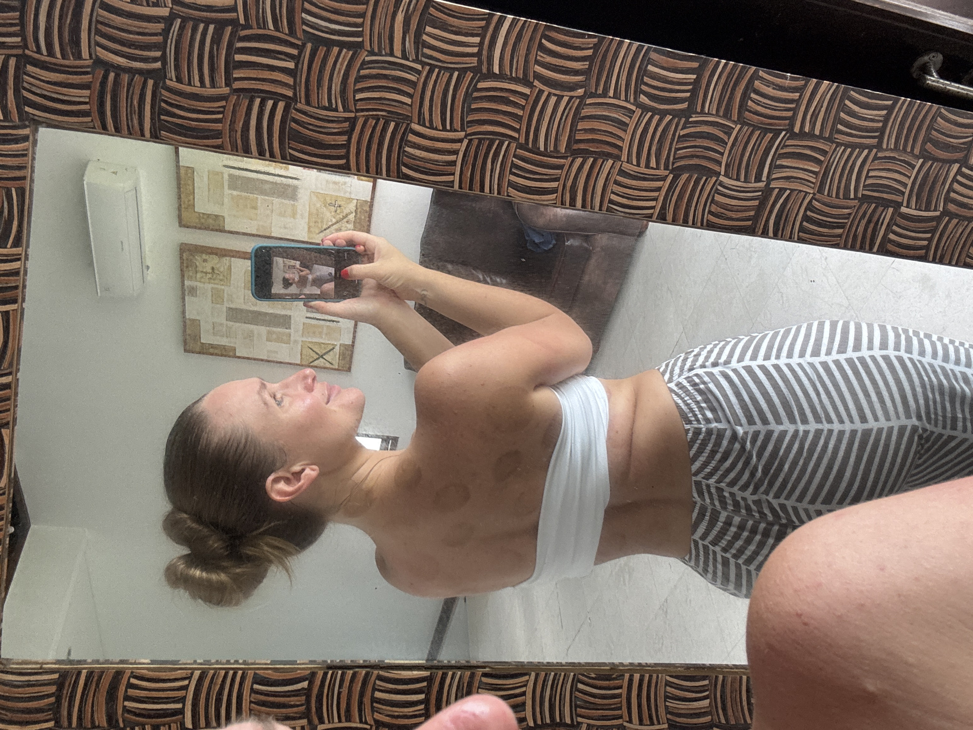 A woman is taking a picture of herself in a mirror. The mirror has a patterned, wooden frame. The woman is wearing a white top and patterned pants. The location appears to be indoors, possibly a bathroom or dressing area, with artwork or decor on the walls. The location in the city of Kuta Selatan, Indonesia cannot be ascertained from the image.