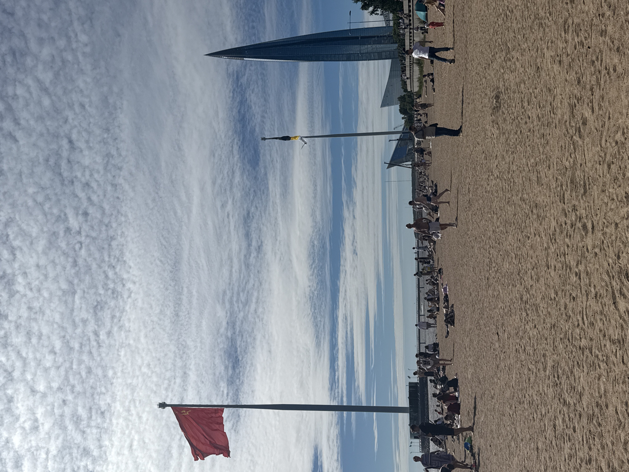 The image depicts a sunny day at a sandy beach in St. Petersburg, Russia. A large, red flag flies from a tall flagpole on the left side of the frame. In the background, a distinctive, modern glass skyscraper with a pointed top dominates the skyline. Another flag, yellow and black, is visible on a second flagpole. The beach is populated with numerous people, some lying on towels, others standing or walking. The sand is textured, and the sky is filled with scattered white clouds. The water of the Gulf of Finland is visible in the distance. The scene captures a leisure activity on a pleasant day.