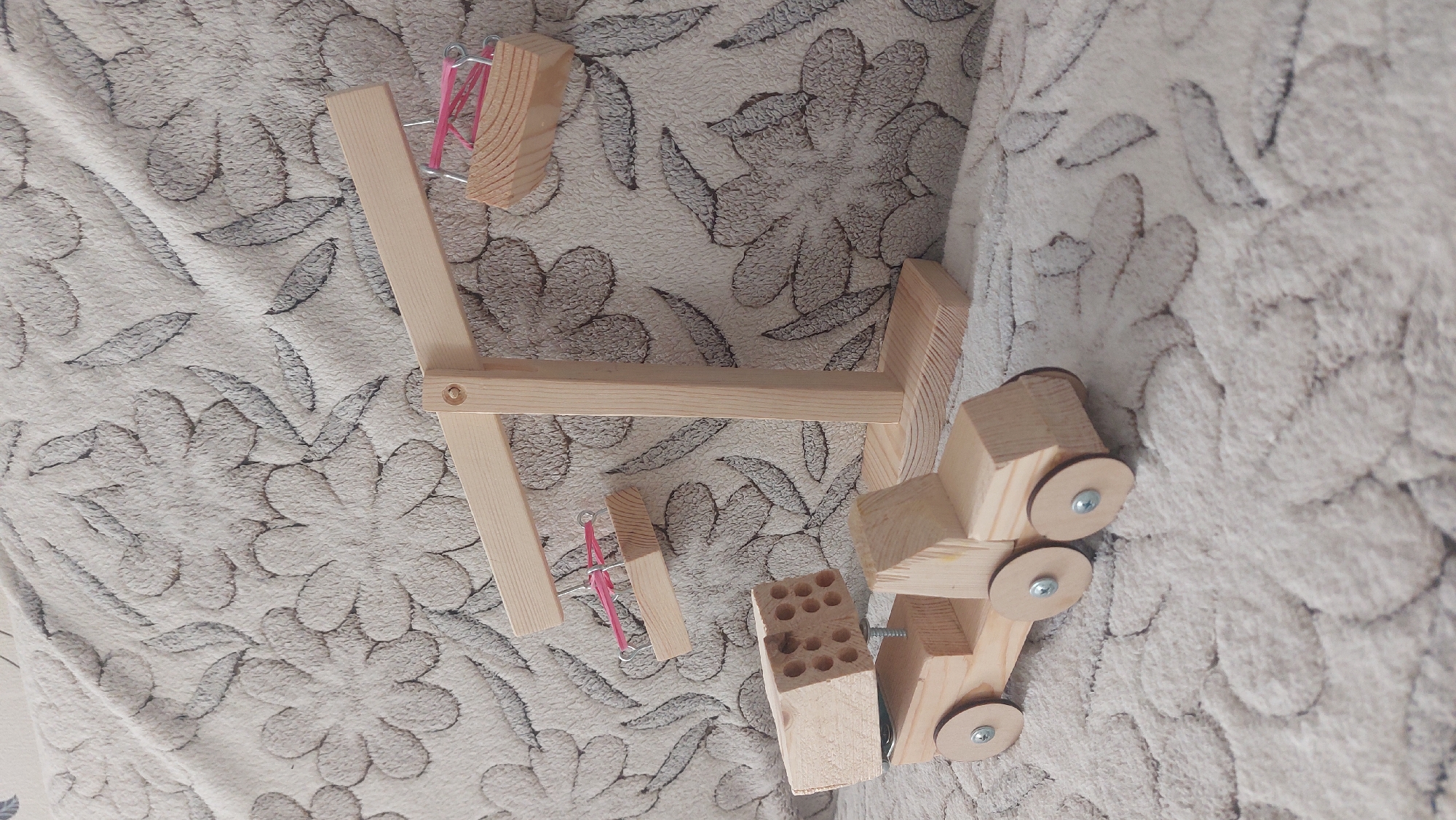 The image shows a collection of handcrafted wooden toys arranged on a textured fabric surface with a floral pattern. The toys include a vehicle with wheels attached by screws, a block with drilled holes, and a wooden A-frame structure, parts of which are held together with rubber bands and metal staples. The setting is unidentifiable, there is nothing within the image to confirm this image was taken in Novosibirsk, Russia.