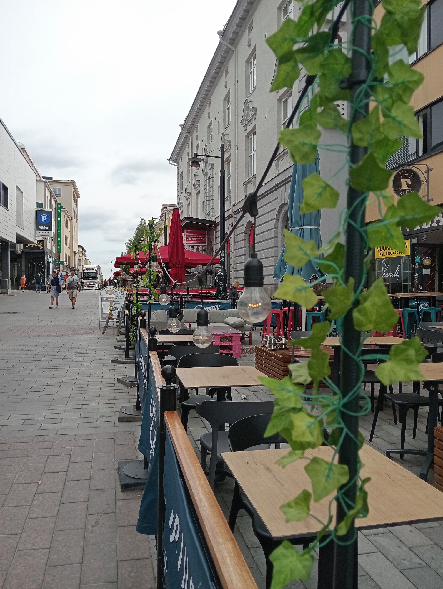 The image depicts a street scene in the city of Kuopio, Finland, likely on or near Puijonkatu. The foreground features an outdoor seating area of a cafe or restaurant, characterized by wooden tables, black chairs, and dark blue banners. A black pole adorned with artificial green ivy and string lights runs vertically through the frame. Beyond this seating area, the sidewalk is paved with grey interlocking bricks.
Several people are visible walking on the sidewalk. Two men, one wearing a grey shirt and the other a blue shirt, are walking towards the camera. Further down the street, a woman in an orange dress is walking away from the camera. In the background, a large truck is visible, as are various buildings lining the street, some featuring storefronts. Red umbrellas for outdoor seating are also prominent. The sky is overcast.