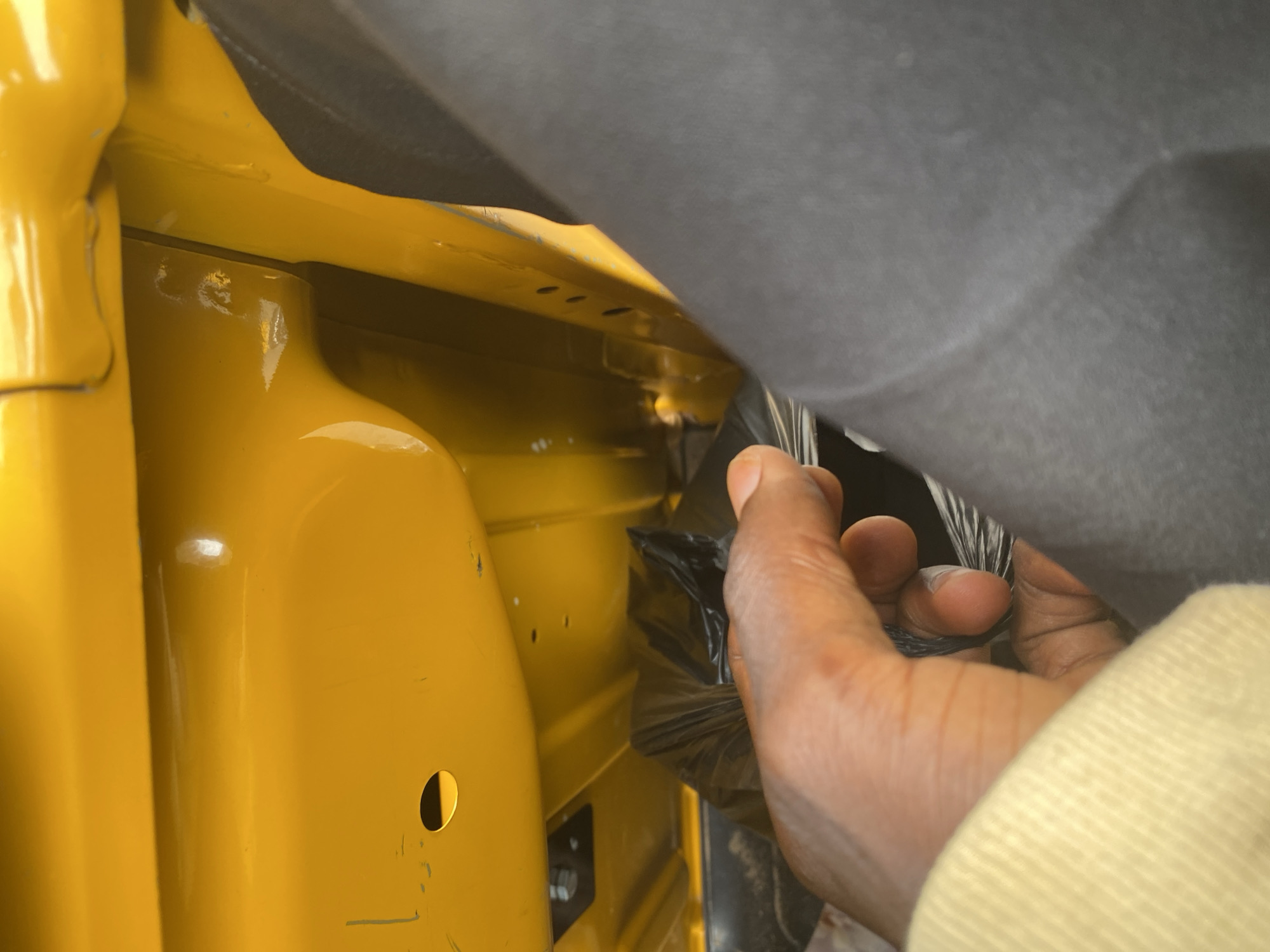A close-up shot shows a person's hand reaching into a yellow compartment, possibly the interior of a vehicle. The hand, with dark skin, is holding a bundle of black plastic material. The compartment is painted a bright yellow and appears to have a ribbed texture in places. Above the yellow compartment, a dark gray material, possibly a fabric cover, is draped, obscuring some of the space. The lighting suggests it is daytime, with natural light illuminating the scene. The background is slightly blurred, focusing attention on the hand and the yellow compartment. The presence of the plastic material being held suggests an action of insertion, removal, or organization within the compartment. The overall impression is one of a functional space being accessed or adjusted. The location context of Jalingo, Nigeria, implies an outdoor or semi-outdoor setting, possibly related to transport or storage.